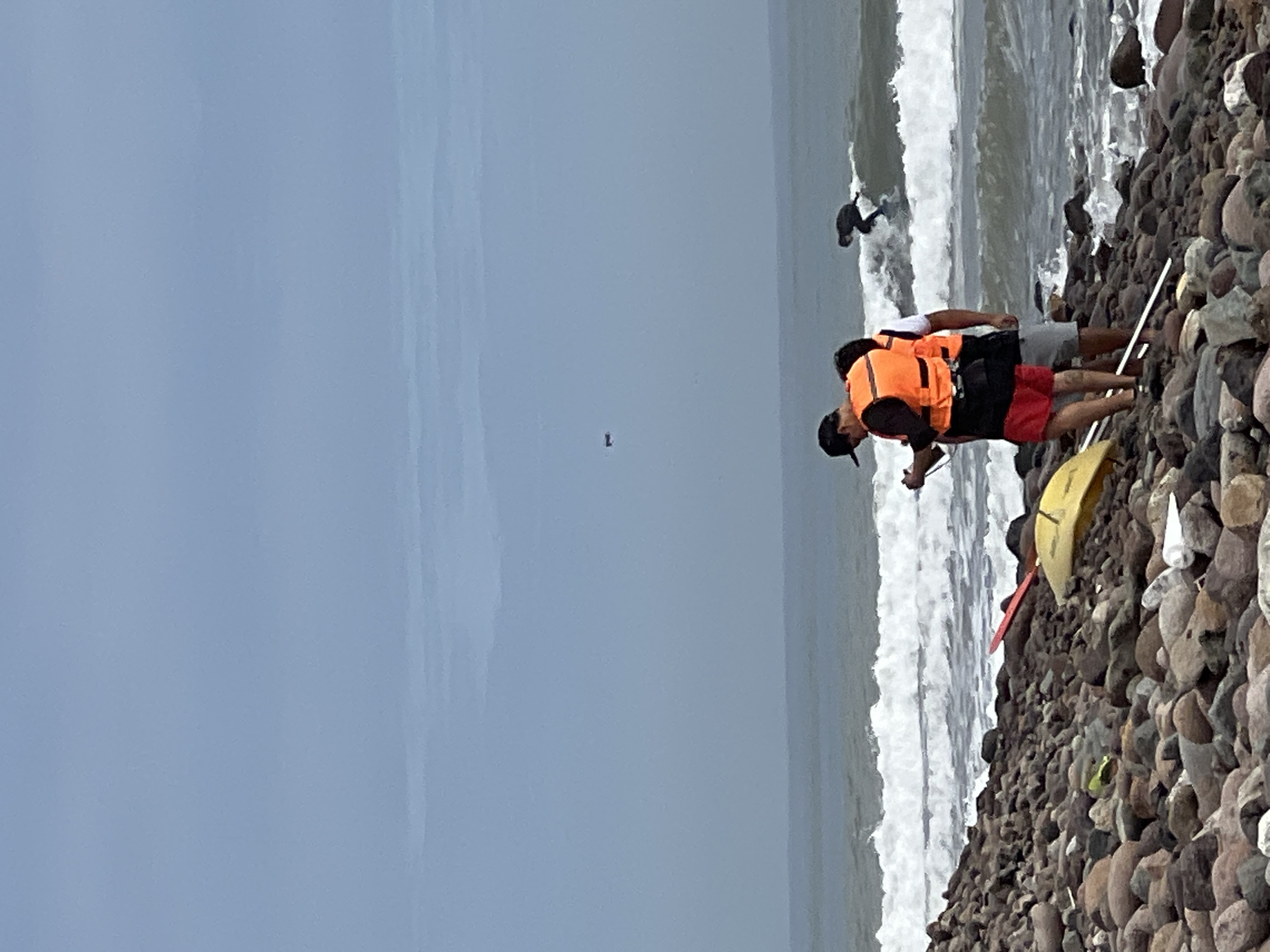 The scene depicts a rocky beach in Arica, Chile, with a person surfing in the waves and two people standing on the shore, one wearing an orange life vest. A yellow kayak rests on the rocky beach. The sky is overcast.