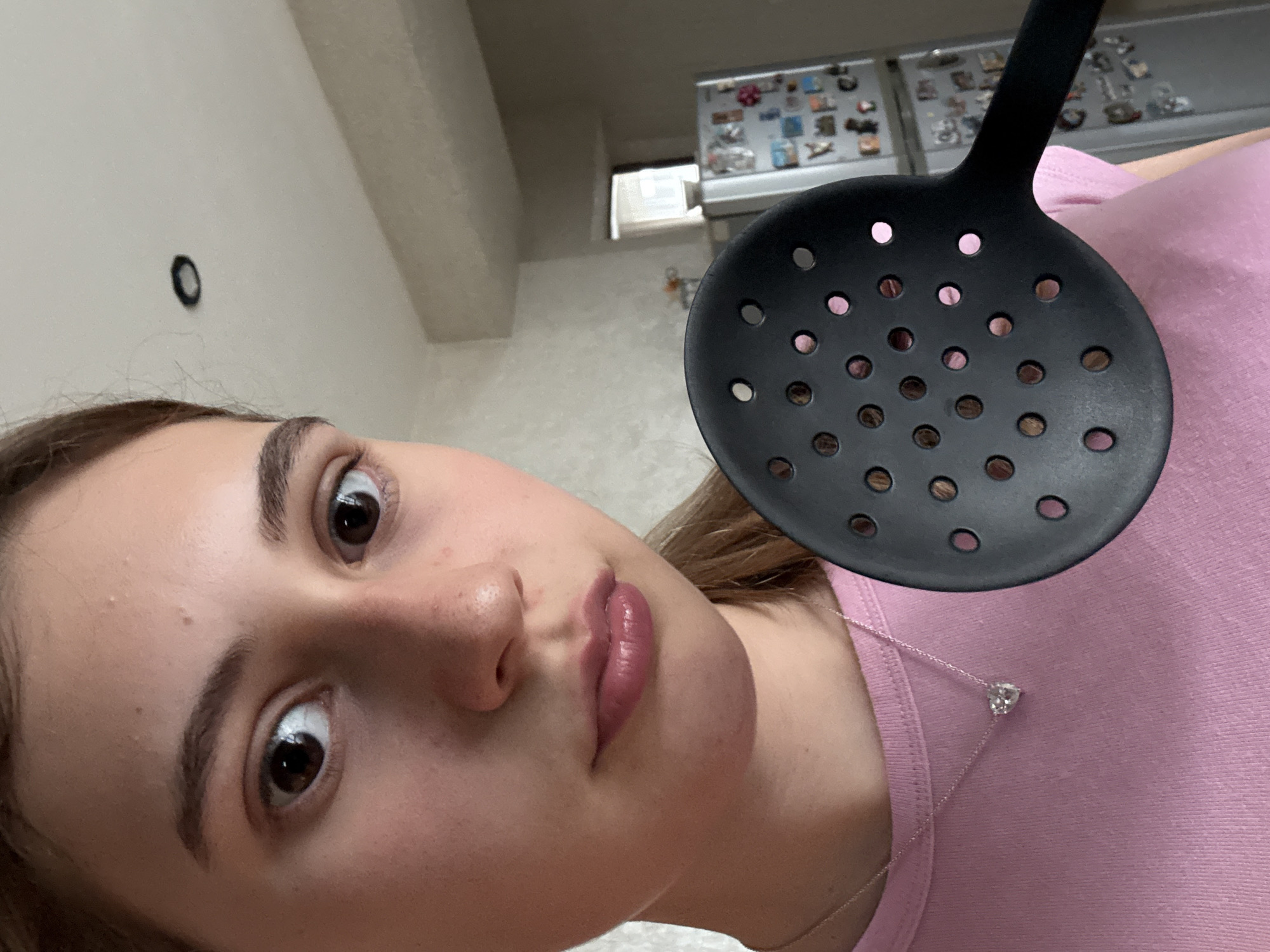 A close-up, tilted selfie shows a young woman holding a black plastic slotted spoon near her face. She has brown eyes and light brown hair, wears a pink shirt, and a silver necklace with a heart-shaped pendant. In the background, blurred, is part of a kitchen with a refrigerator visible, which is decorated with magnets. The location is Panagyurishte, Bulgaria.
