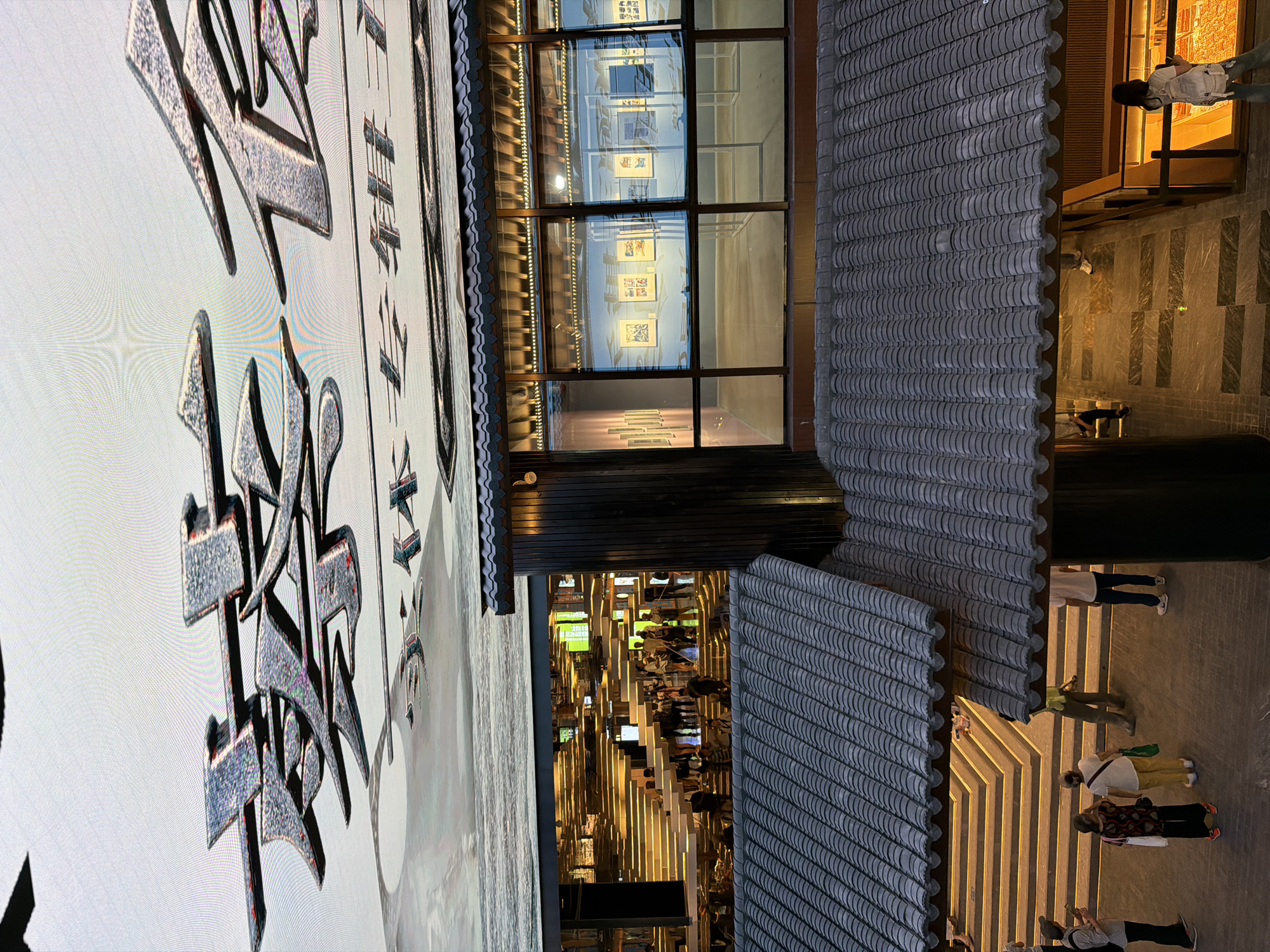 This image depicts an outdoor scene with a mix of traditional and modern architectural elements in the Yuzhong District, China. In the foreground, two traditional tiled roofs are prominently featured, with one extending over a walkway. Below these roofs, a modern building with large glass windows is visible, revealing an art exhibition inside. Through the windows, several framed artworks are displayed on the walls. Beyond the exhibition space, a well-lit interior with tiered seating, resembling a grand staircase, is visible, with a number of people standing and walking.
In the outdoor area, several individuals are present. Near the tiered seating, a group of people are gathered, some looking towards the interior. Further down, closer to the foreground, three women and two men are standing on a paved area. One woman in the foreground is wearing a black and red patterned top and dark pants, holding a white bag. Another woman next to her is wearing a white top and yellow pants, carrying a green bag. A man in a white t-shirt and blue jeans stands to the right, while another man in a lime green shirt and dark pants is further in the background. A woman on the far left is wearing a hat and carrying a bag.
Above the scene, a large digital display or screen is visible, featuring what appear to be stylized Chinese characters in a metallic texture, with a reflective surface below them. The overall atmosphere suggests an evening or late afternoon setting, given the artificial lighting illuminating the interior spaces and the screen.