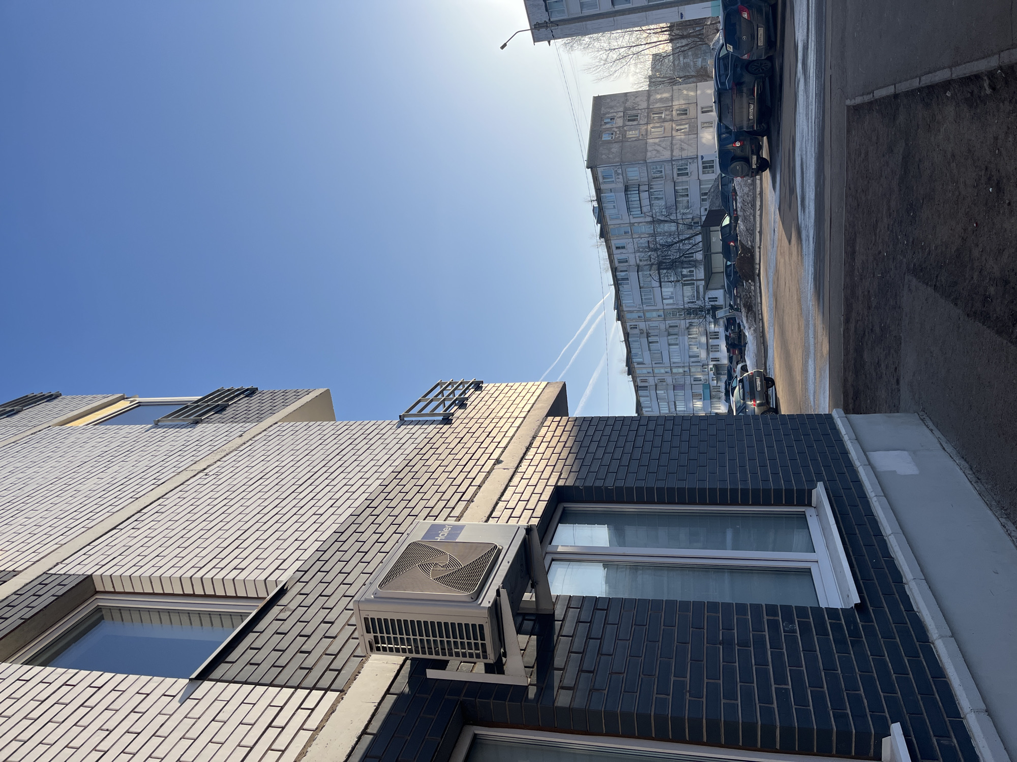 The image shows an upward shot of a building in what is likely the city of Chaikovsky, Russia. The building features a mix of white and dark brickwork, with an air conditioning unit mounted on the facade. Windows are visible. In the background, there is an older, multi-story building, along with parked cars and a sidewalk. The sky is clear blue, indicating a sunny day. There are contrails visible in the sky, likely from an airplane. The angle of the photo gives a ground-level perspective, and the details suggest an urban environment.