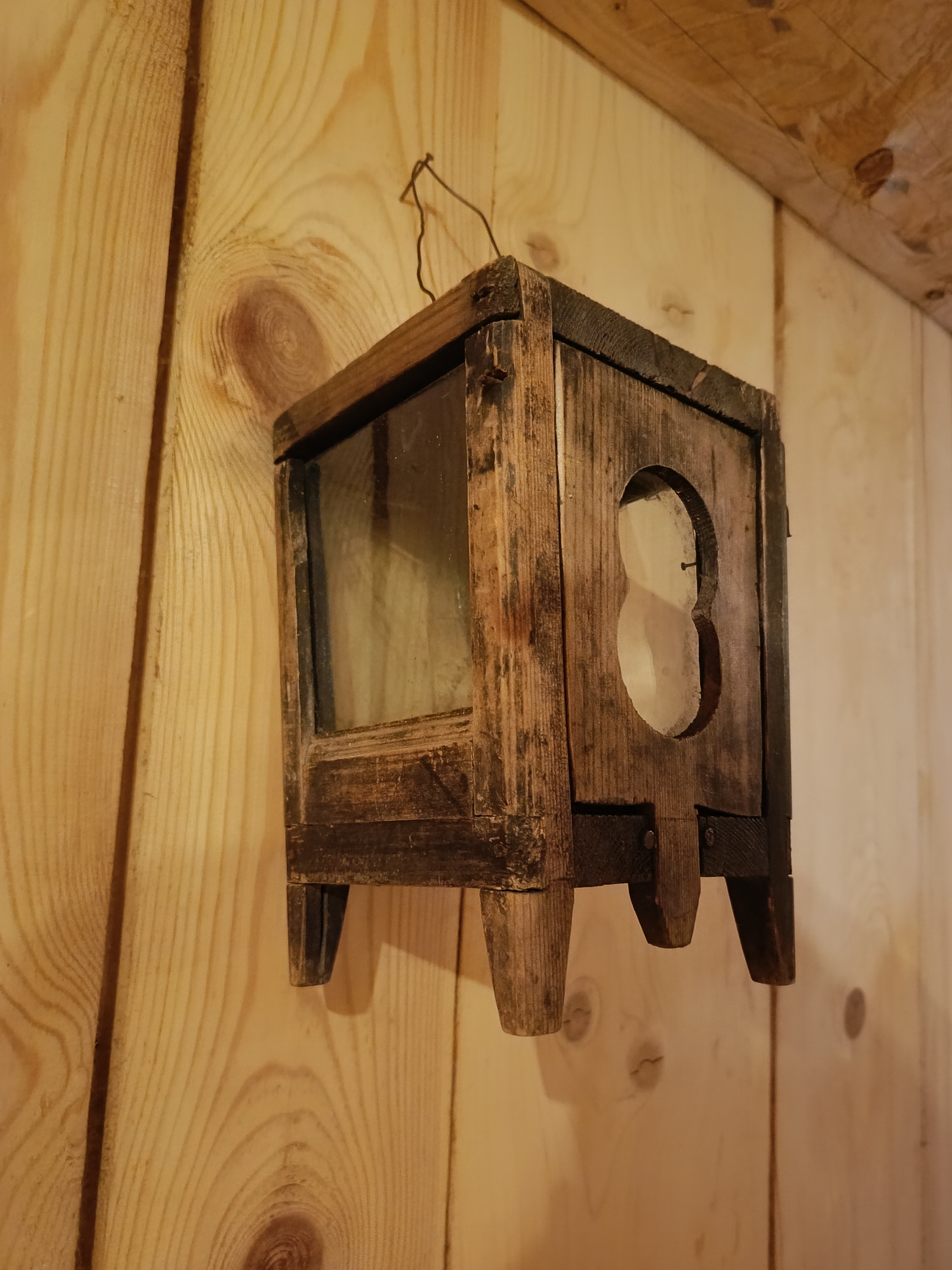 The image shows a weathered wooden object, possibly a birdhouse or lantern, hanging on a light-colored wooden wall. The object is square-shaped with a glass panel on one side and a rounded cutout on another. It has four short, tapered legs. The wall is made of vertical wooden planks with visible grain patterns. The lighting suggests an indoor environment, possibly a rustic setting like a cabin or workshop. The object is suspended by a thin wire attached to its top. The overall impression is one of age and handmade construction.