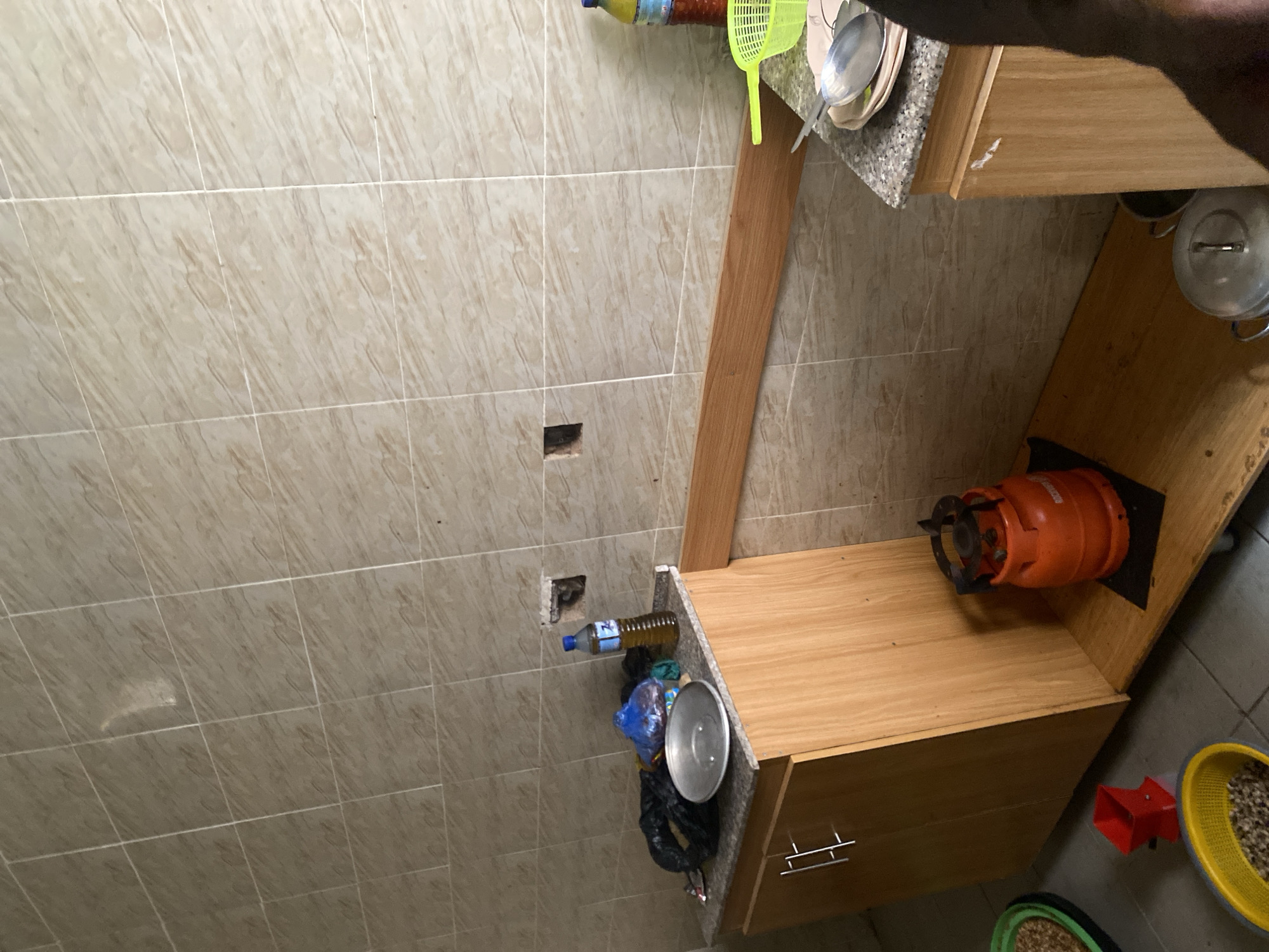 The image shows a cluttered kitchen counter and surrounding area in Dumnga, Nigeria. The scene is characterized by a beige tiled wall, a wooden cabinet, and a granite countertop. On the countertop, there are several objects, including a bottle of cooking oil, a green fly swatter, and a stack of plates with a spoon. Next to these, a gas cylinder is visible, connected to a burner. Various items are piled on the counter, including trash bags, a metallic lid, and a plastic container. In the background, a large metal pot is partially visible on a shelf. The floor is tiled, and a yellow basket filled with what appears to be grains is placed on the floor, along with other containers. The lighting suggests it is daytime. The overall impression is one of a busy and functional, albeit somewhat disorganized, kitchen space. There is no visible text in the image.