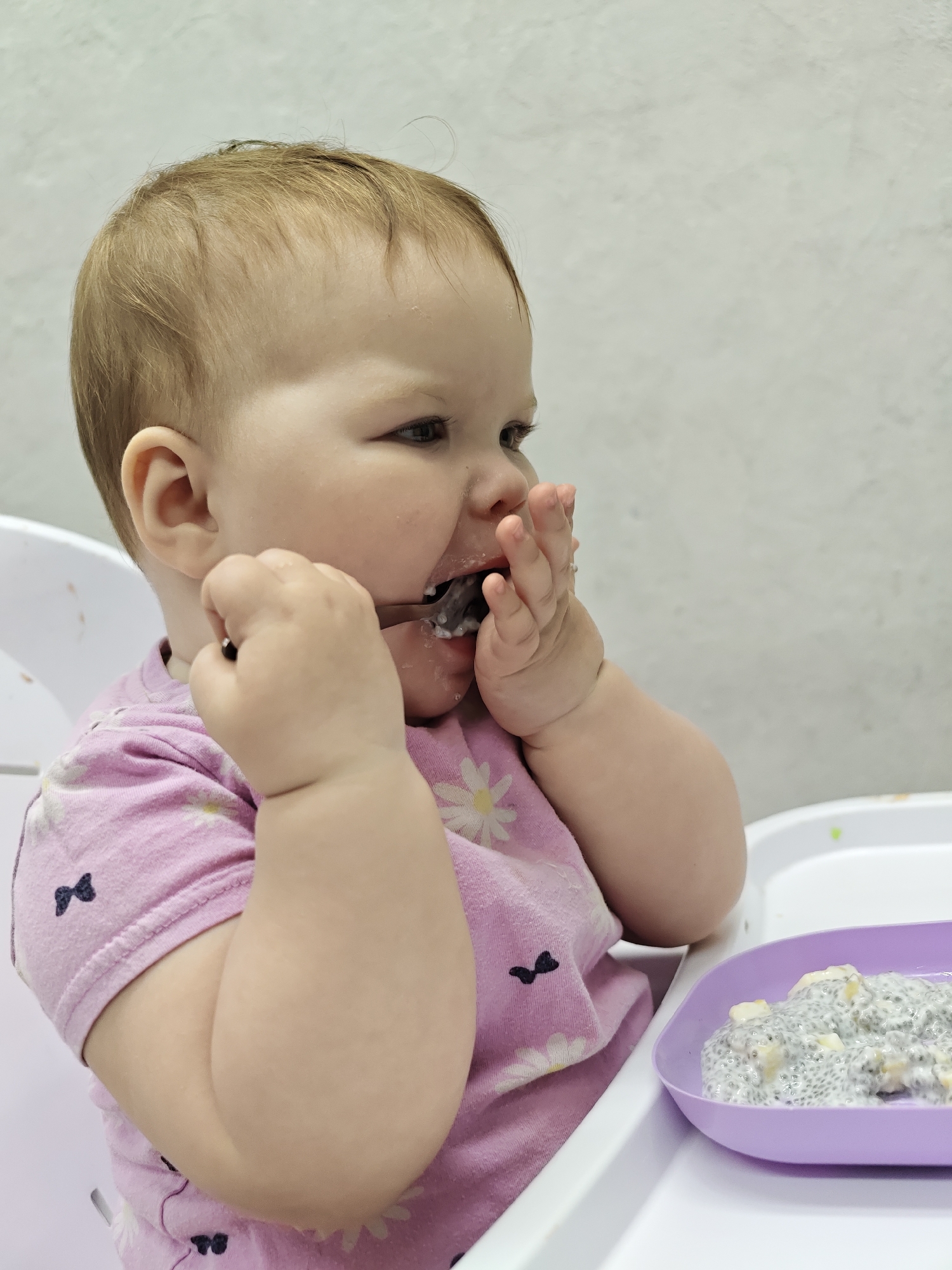 A baby sits in a high chair, positioned on the right side of the frame. The baby is fair-skinned with light brown hair and is wearing a light pink short-sleeved shirt with a daisy and bow pattern. The baby's hands are near their mouth, one holding a spoon, and the other near their face. There appears to be food on the baby's face and hands. A purple plate with a white and grey mixture, likely the baby's food, rests on the high chair's tray. The background is a plain, light-colored wall. No specific evidence suggests the image was taken in Chaikovsky, Russia.