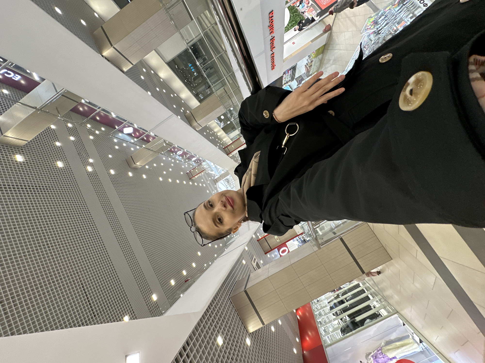 A low-angle, tilted selfie captures a woman inside a multi-level shopping mall. The woman, who appears to be the photographer, is looking towards the camera with a neutral to slightly smiling expression. She wears dark-rimmed glasses perched on her nose and is dressed in a black coat with large, light-colored, patterned buttons visible on the lapel and sleeve. A metallic, circular keyring or latch hangs from her coat. Her left hand is partially visible, resting near her chest.
The background reveals the modern interior architecture of the shopping center. Multiple levels of the mall are visible, characterized by white walls, glass railings, and metallic structural elements. The ceiling features a distinctive grey grid pattern with numerous small, circular embedded lights, creating a bright, evenly lit environment. Below, glimpses of storefronts and escalators can be seen. On an upper level, a red sign with white text is partially visible, reading "красота · ход забота" (krasota · khod zabota), which translates to "beauty · care course" or "beauty · concern/care process". Another partially obscured sign shows "ECI". The setting suggests an active shopping area, likely during the daytime given the ample indoor lighting. The overall impression is a bustling, contemporary commercial space in Yekaterinburg, Russia.