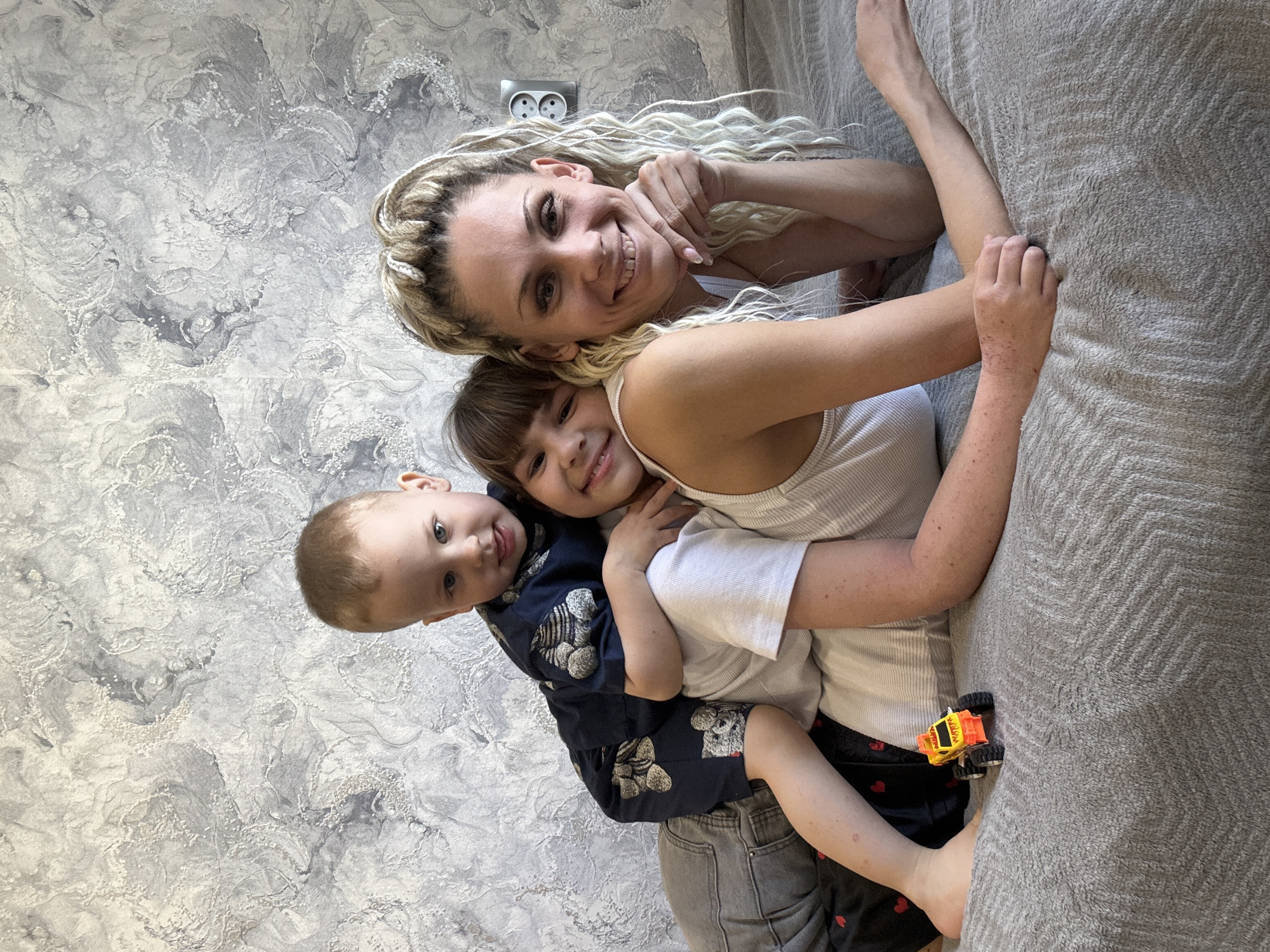 A woman with blonde braided hair is lying on a gray bedspread, smiling, with two children. The woman is wearing a white tank top. One child, who appears to be a toddler, is wearing a navy blue outfit with teddy bear prints. The other child, appearing to be older, is wearing a white shirt. The children are close to the woman. The background consists of a gray textured wall with a white electrical outlet visible. A yellow and orange toy truck is near the woman's arm on the bed. There are no identifiable elements indicating the location of Заволжье, Russia.
