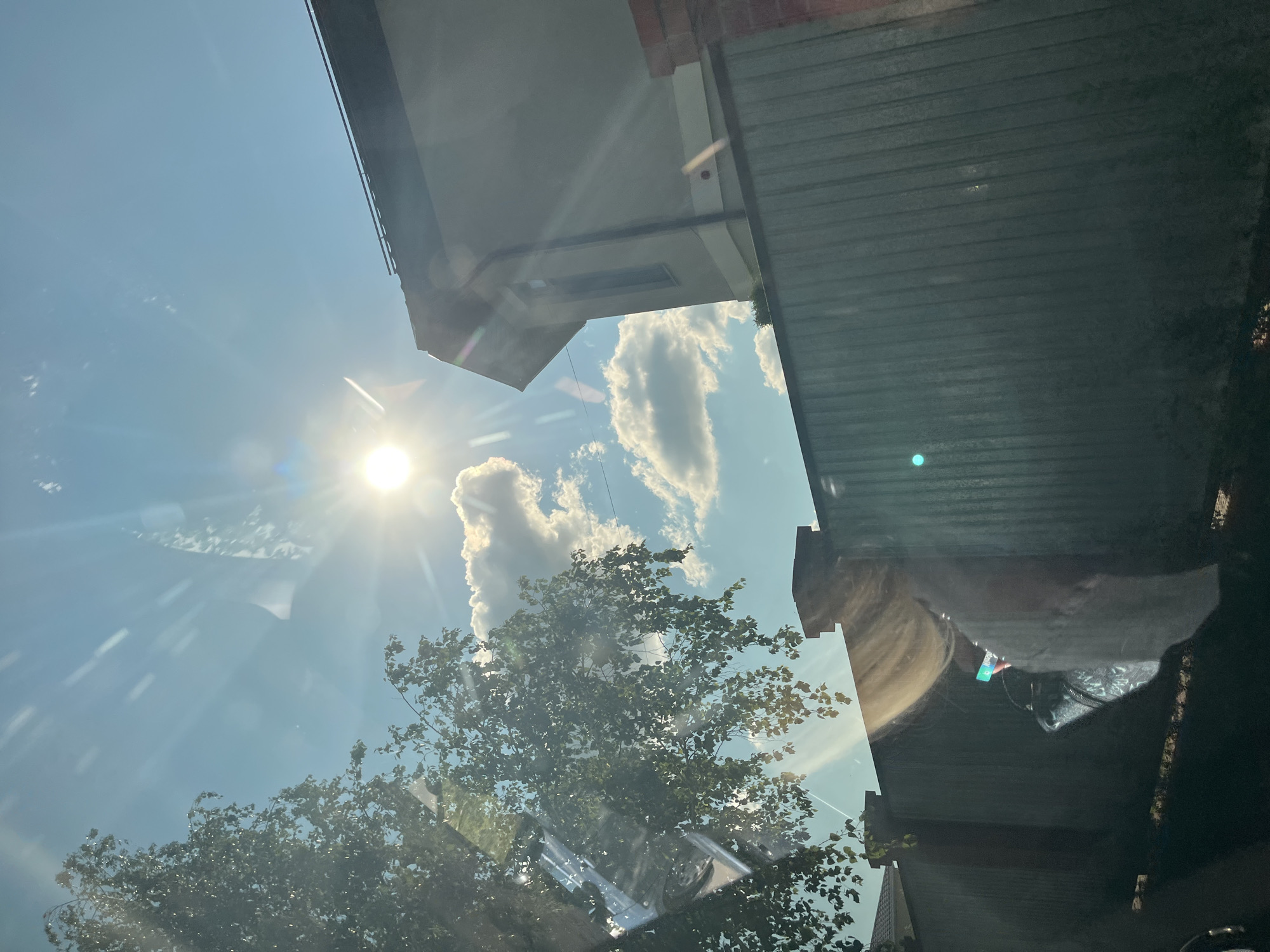 The image depicts a sunny day with bright sunlight shining through a tree in the foreground. The sun is near the center of the frame, with rays extending outwards. In the background, there is a light-colored house with a dark roof and a light gray metal fence. Several clouds are visible in the blue sky. Reflections are present on the surface from which the photo was taken, showing a person with blonde hair holding a phone. The scene appears to be in a residential area.
