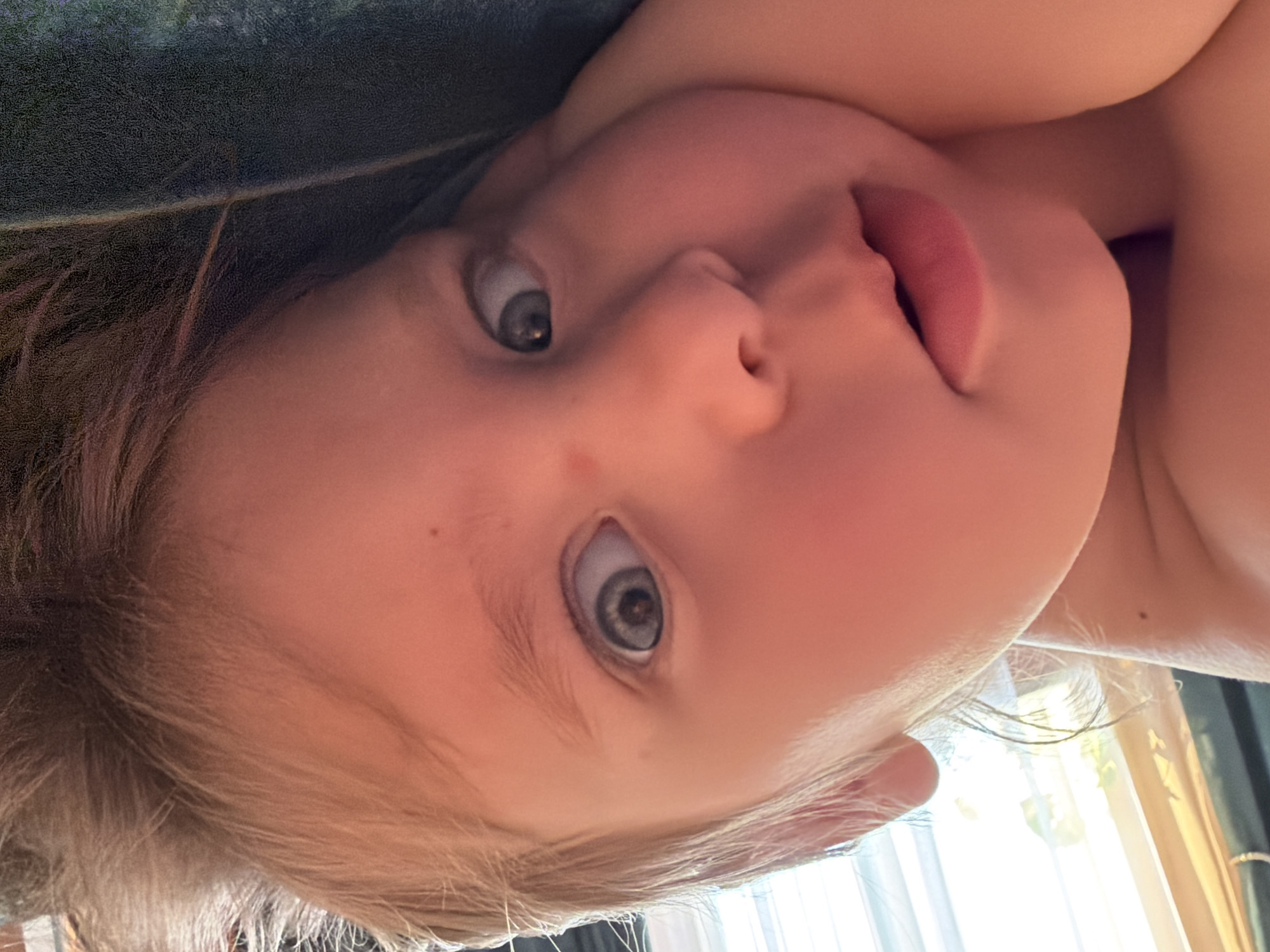 A close-up, upward-facing perspective captures a young child with light blonde hair and fair skin. The child has light-colored eyes and slightly parted lips. The background is partially obscured, but a textured dark surface is visible at the top left, and a bright window with a curtain is at the bottom right. There is no visible evidence to link this image to Chaikovsky, Russia. The subject of the image is a child looking up at the camera. The environment appears to be indoors.