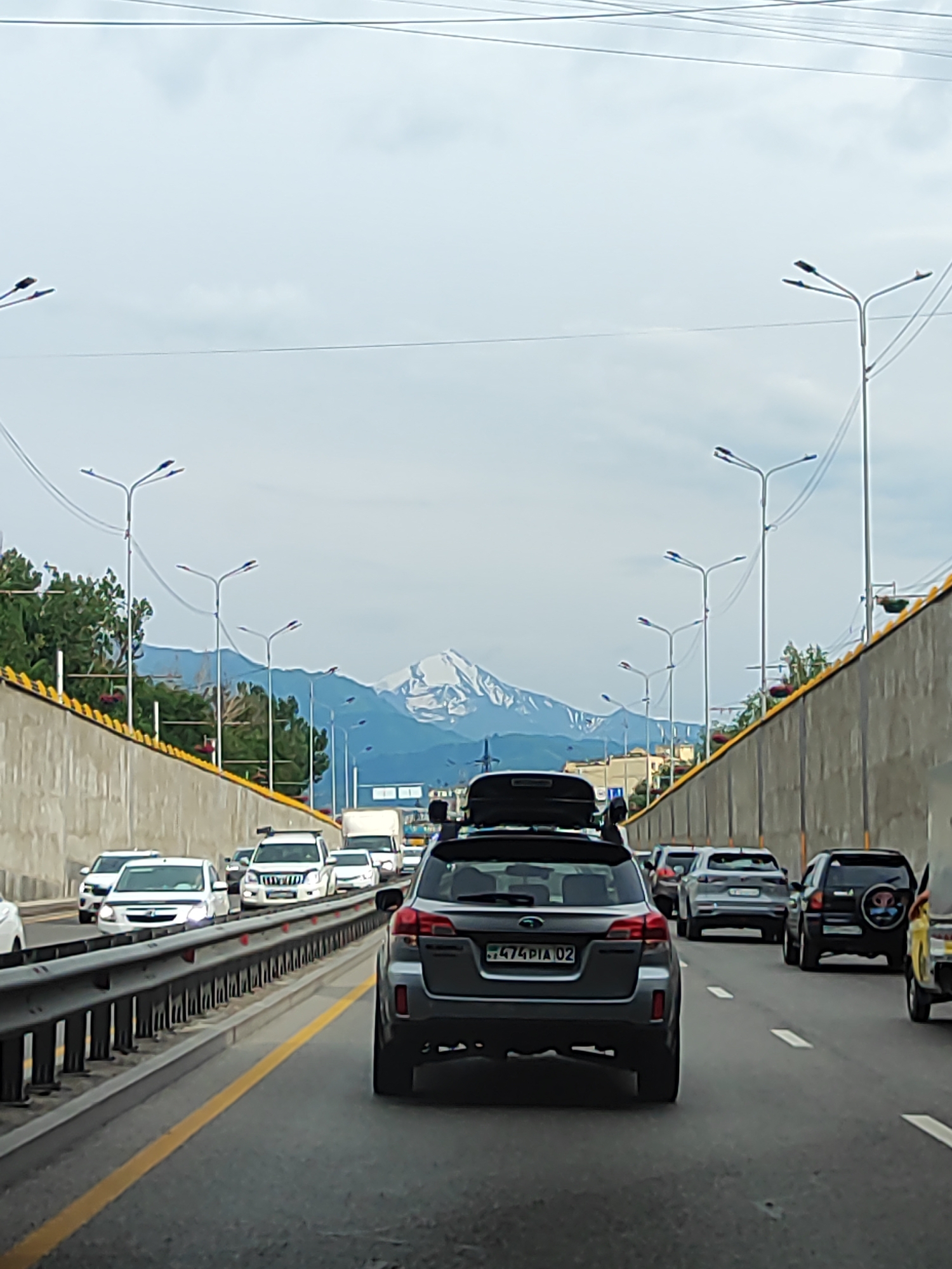 This media file shows a busy highway in Almaty, Kazakhstan, with a traffic jam under a cloudy sky. The foreground features the rear of a dark gray SUV, possibly a Subaru Outback, with a roof-top cargo carrier and a license plate reading "474 DIA 02". Other vehicles are visible in the traffic, including a white van, a black SUV with a spare tire on the back, and several other cars.
The highway has multiple lanes, separated by a yellow line and a metal guardrail. On the left side, a concrete wall topped with yellow and green accents runs alongside the road, with trees and streetlights visible above it. On the right side, a similar concrete wall borders the highway, with more streetlights and some buildings in the distance.
In the background, a majestic snow-capped mountain range dominates the skyline, suggesting a location near the Tien Shan mountains, a prominent geographical feature of Almaty. The weather appears overcast, with a light, diffused sky. The time of day is likely daytime, given the lighting conditions. The overall scene depicts a typical urban commute, with the natural beauty of the mountains providing a striking backdrop to the everyday hustle of city life. No people are explicitly visible, but the presence of numerous cars implies their presence within the vehicles.