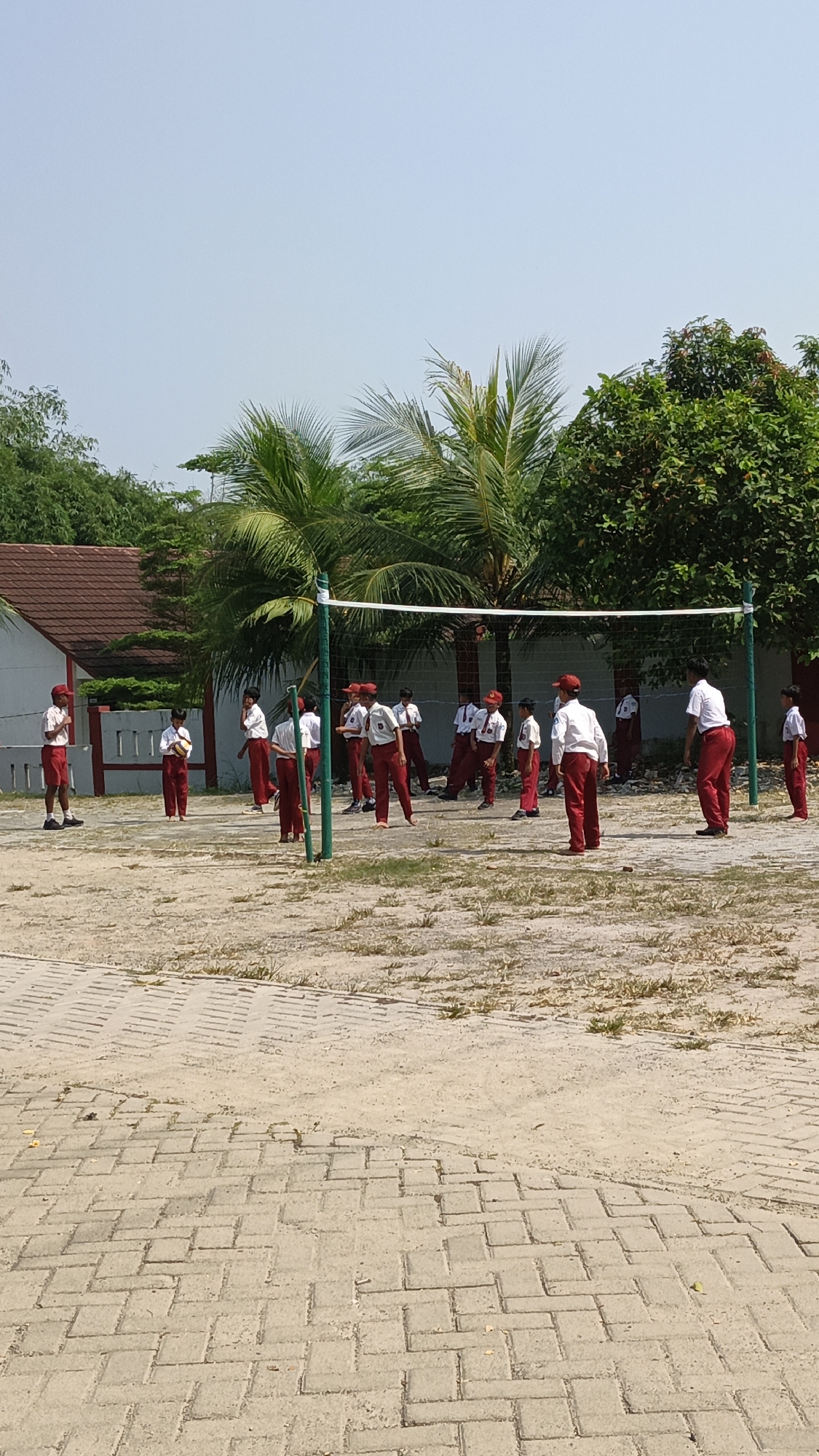 A group of schoolchildren in red shorts and white shirts with red caps are playing volleyball on a sandy court in Pasir Bolang, Indonesia. A volleyball net is set up across the court, with players positioned on both sides. Palm trees and other foliage are visible in the background, along with a building with a red roof. The sky is clear and blue.