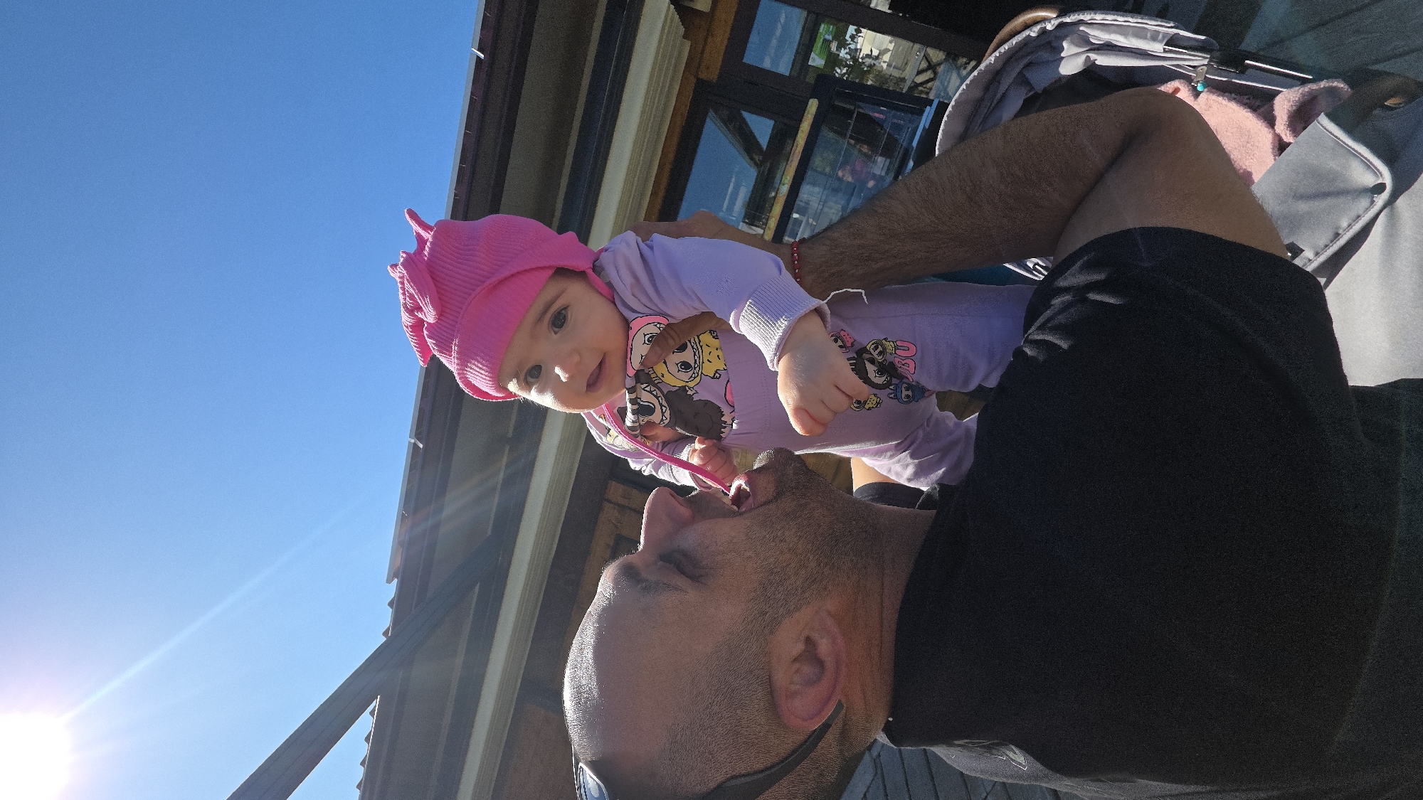 A man holds a baby girl in a pink hat and lavender outfit with Disney characters up to his mouth. The baby has a pink plastic spoon in her hands. The location appears to be outdoors in Varna, Bulgaria, based on the typical architectural elements of the buildings visible in the background. There is a stroller in the upper right. The sky is a clear blue.