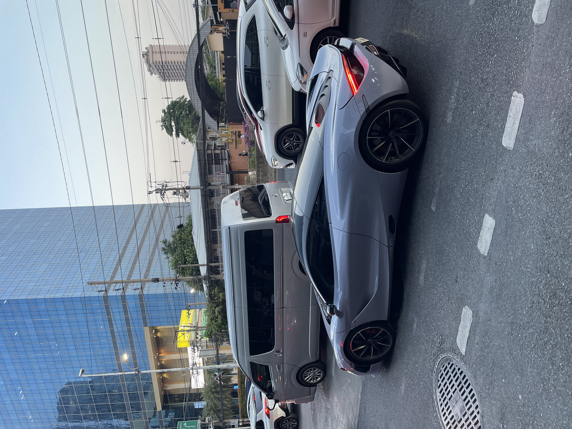 This photograph captures a bustling street scene in an urban environment, likely Bangkok given the provided context. The image, originally taken in landscape orientation, is presented rotated 90 degrees clockwise.
In the immediate foreground, a sleek, light grey sports car, possibly a McLaren, is visible from its rear-right side, with its illuminated red taillights prominent. It features distinctive multi-spoke black wheels with red brake calipers. Directly behind the sports car is a white Mercedes-Benz SUV or crossover, identified by "AMG Line" text on its side trim. Further back in the same lane, a grey passenger van follows, with part of another white vehicle visible behind it. The vehicles appear to be in traffic, suggesting a busy period or a slowdown on the road.
To the left, a modern glass skyscraper with blue-tinted windows reflects the sky and surroundings, dominating the urban skyline. In the distance, an older, lighter-colored building with numerous balconies stands against a pale, hazy sky. The scene is crisscrossed by a dense network of overhead utility wires and poles, a common sight in many Southeast Asian cities.
The time of day appears to be late afternoon or early evening, with soft, diffused lighting and a slightly overcast or hazy sky. The asphalt road surface shows faded white lane markings and a circular metal manhole cover near the bottom right. While individual people are not clearly distinguishable, the setting suggests a lively urban environment. The only clearly readable text is "AMG Line" on the white SUV.