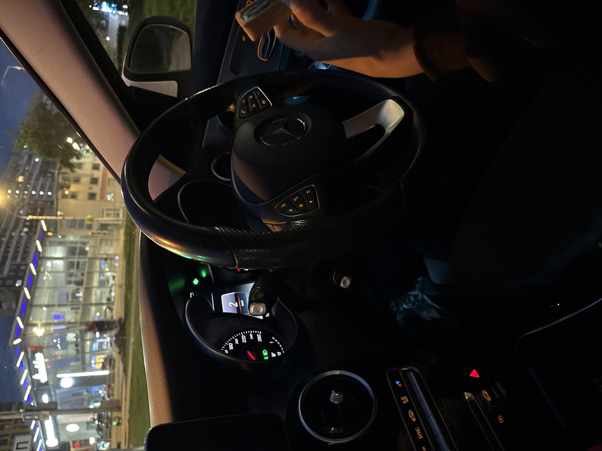 The image shows the interior of a Mercedes-Benz car at night. The car is located in Krasnoyarsk, Russia, judging by the buildings visible through the windshield. The dashboard is visible, displaying a speedometer reading of 2 km/h. A person's hand is visible above the steering wheel, holding an object, possibly a phone or wallet. The vehicle is stationary, as indicated by the speedometer. The scene depicts a combination of artificial lights from the city and the vehicle's dashboard.