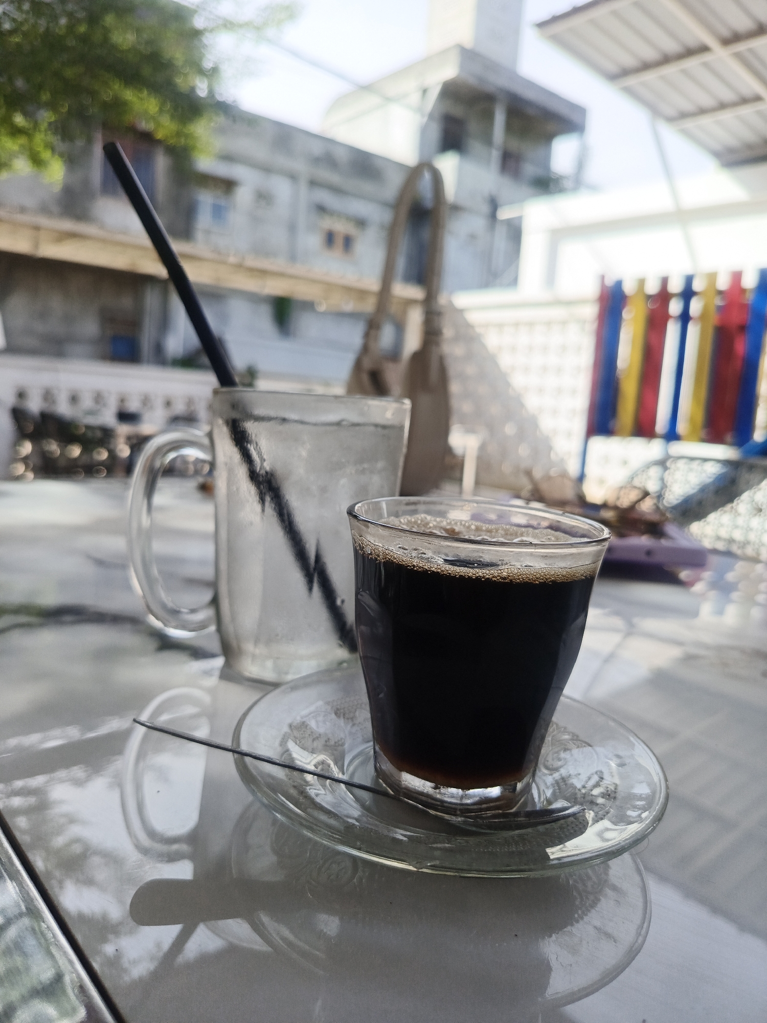The image depicts an outdoor table scene in the city of Pantegajah, Indonesia. In the foreground, two glass beverages are positioned on a light-colored, reflective table surface. A small, clear glass tumbler on the right contains a dark liquid, presumably coffee, topped with a thin layer of light brown foam or crema. This tumbler rests on a clear glass saucer. To its left, a larger, clear glass mug is filled with ice cubes and water, containing a black plastic straw. A thin, black, rod-like object, likely a stirring stick, lies across the coffee saucer. The table surface reflects the light and surrounding objects, showing subtle variations in tone and texture.
The midground is out of focus but reveals an urban environment. On the left, a portion of a grey building wall with an obscured window and some green foliage is visible. On the right, a structure with vertical colored bars (red, yellow, blue) stands prominently, possibly part of a fence or decorative element. Additional blurry objects, including a light-colored bag, are situated in the background, suggesting other patrons or personal belongings.
The background is heavily blurred, displaying various indistinct architectural structures and a bright, clear sky, consistent with an outdoor setting under daylight. No individuals are present in the frame.