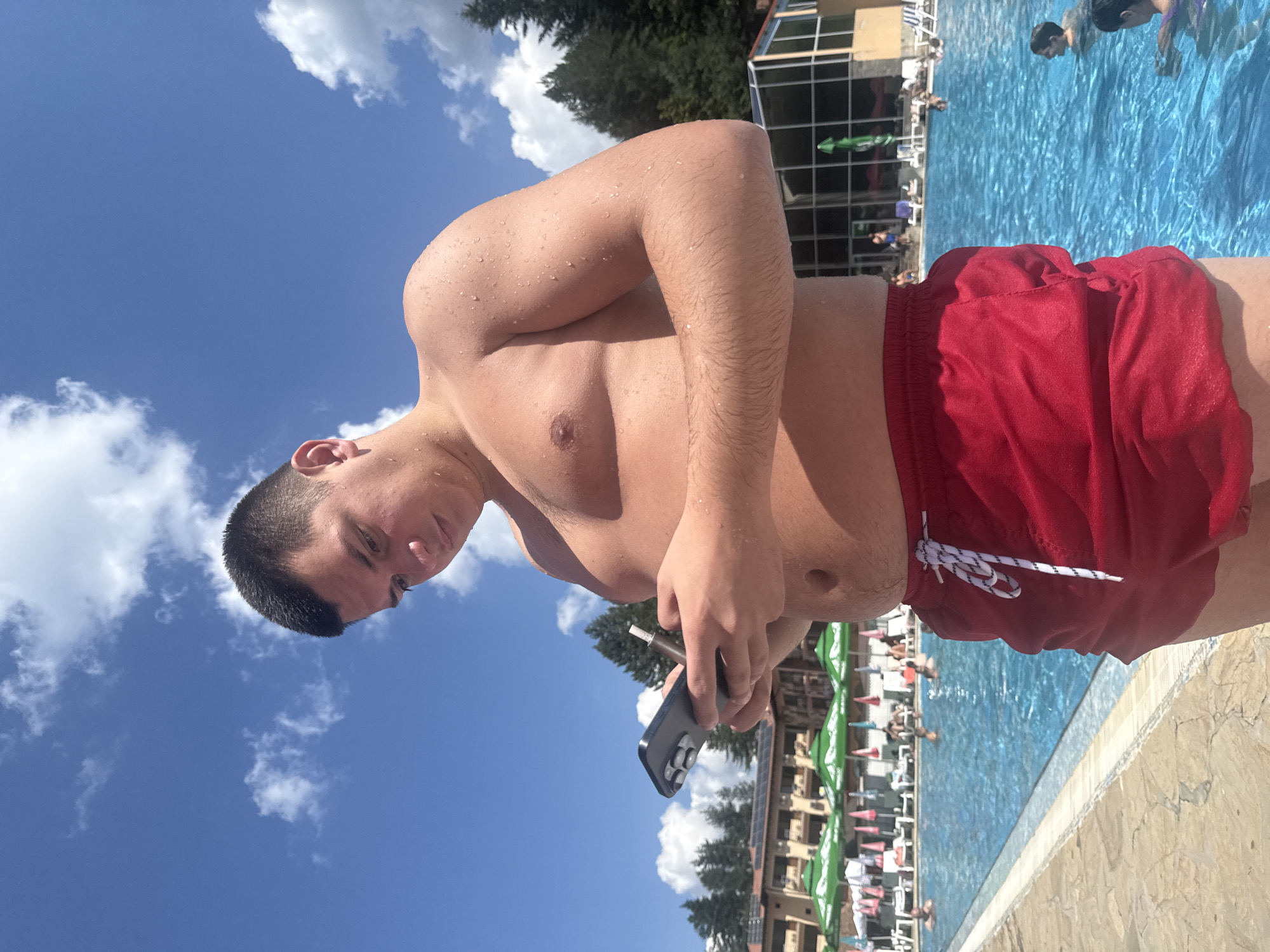 A young man in red swim trunks stands by a swimming pool on a sunny day, looking at his phone and holding a smoking device. He is shirtless and his skin is covered in water droplets. In the background, there are lounge chairs, umbrellas, buildings, and trees. Several other people are visible in the pool. The location is the outdoor swimming pool of a hotel in Chiflik, Bulgaria.