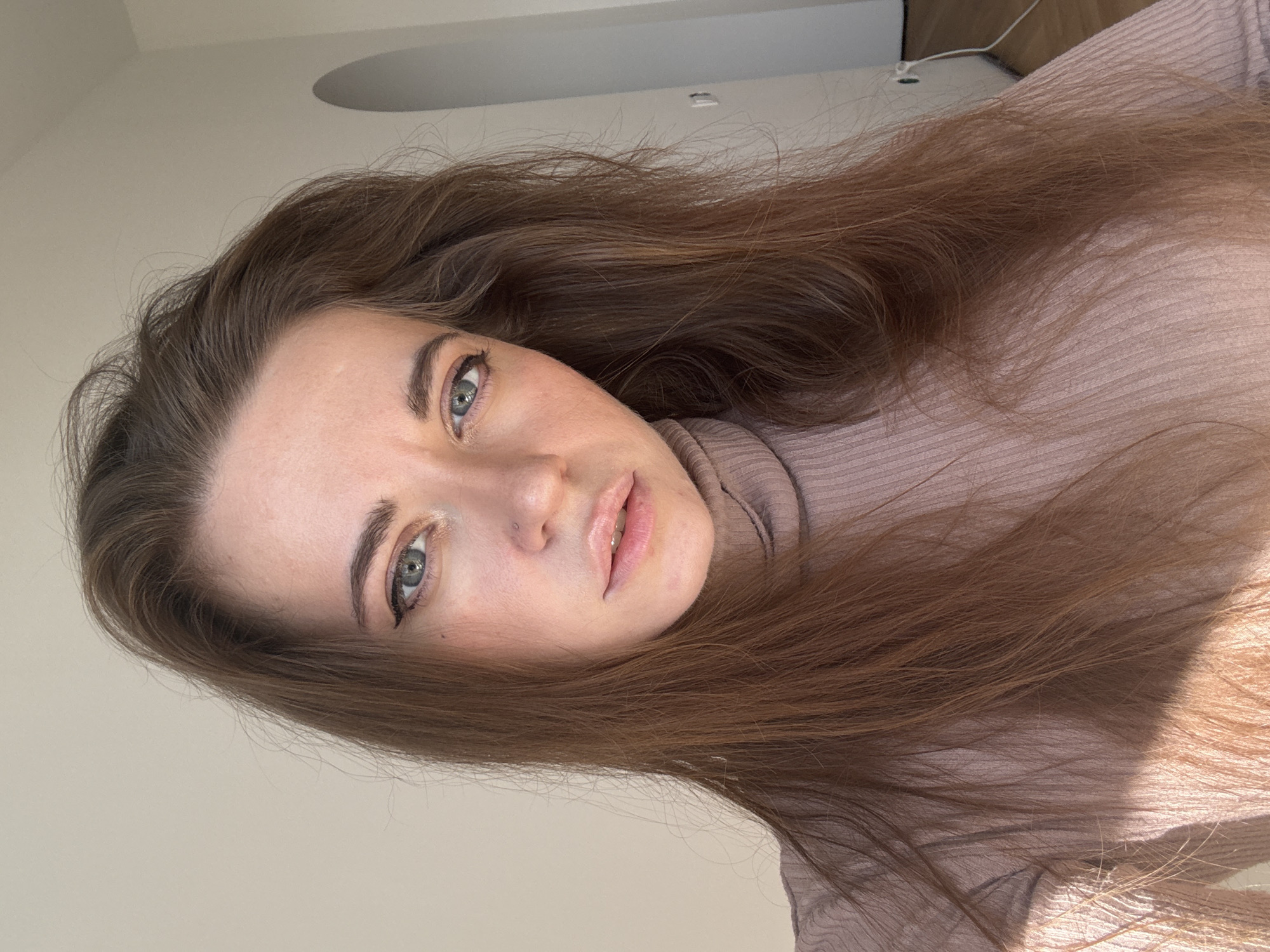 A selfie of a fair-skinned woman with long, brown hair. She is wearing a light brown ribbed turtleneck sweater. The background is a light beige wall with a partially visible ceiling and what appears to be a light fixture. The photo appears to be taken indoors with natural light. There is no evidence to suggest the location is Moscow, Russia.
