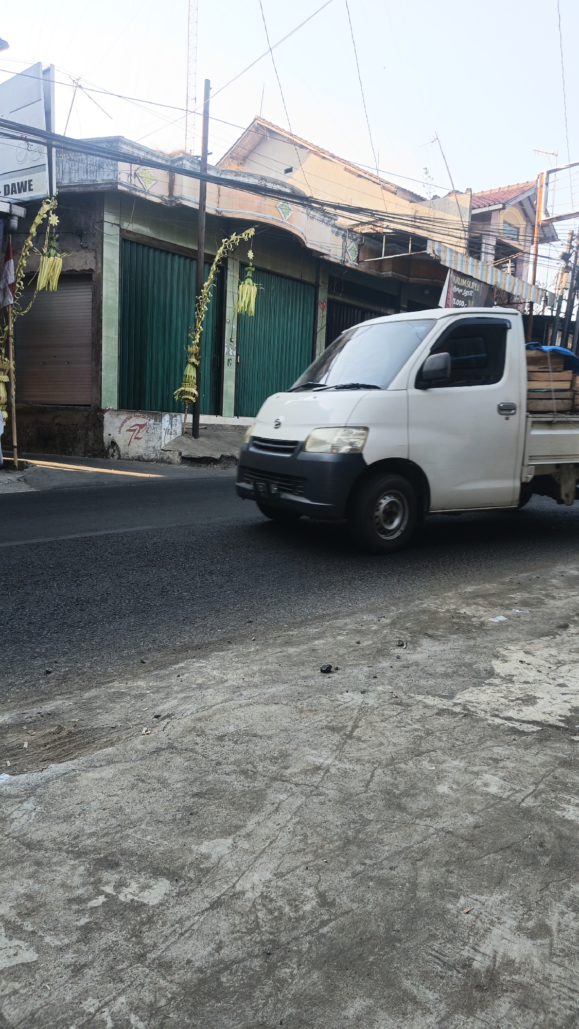 A white utility truck is driving on a paved road in front of a row of shops in Dawe, Indonesia. The shops have green roll-up doors and some are adorned with yellow, leafy decorations. The truck has its headlights on and appears to be moving at a moderate speed. The sky is clear and bright, indicating it is daytime. The foreground of the image shows a rough, concrete surface with some debris.