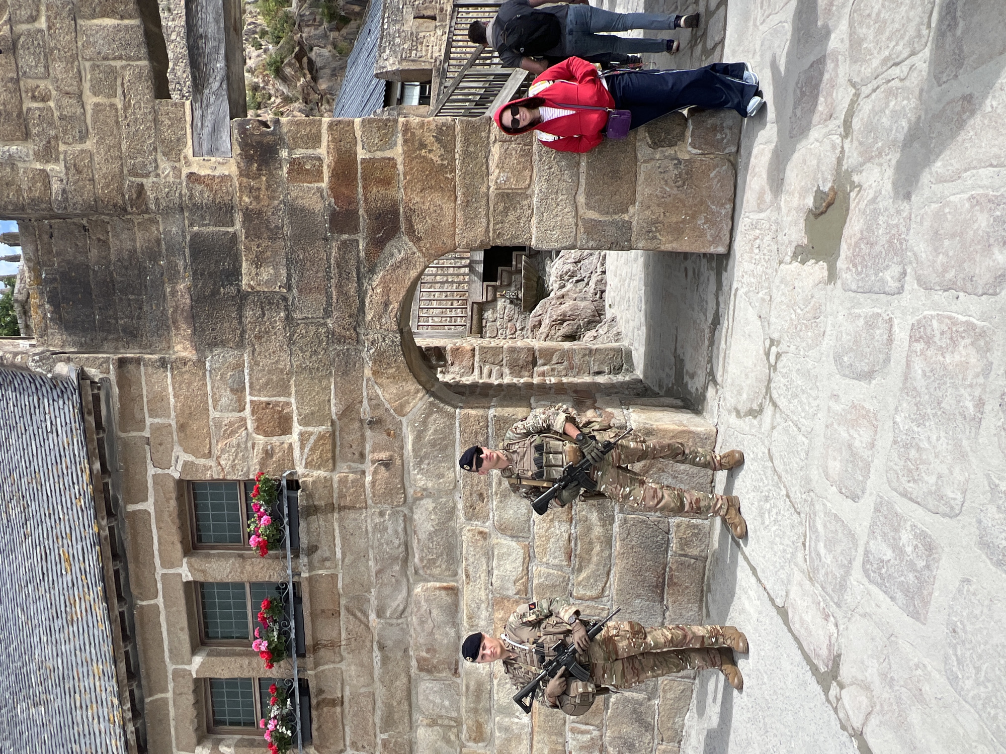 In the city of Le Mont-Saint-Michel, France, two soldiers in camouflage uniforms and black berets stand guard outside a stone building. They are armed with rifles and appear to be positioned at an entrance. To the right of the soldiers, a woman wearing a red hooded jacket and sunglasses leans against a stone pillar. Behind her, a person with a black backpack walks away. The building features stone walls, windows with flower boxes, and an arched doorway. The ground is paved with stone slabs.