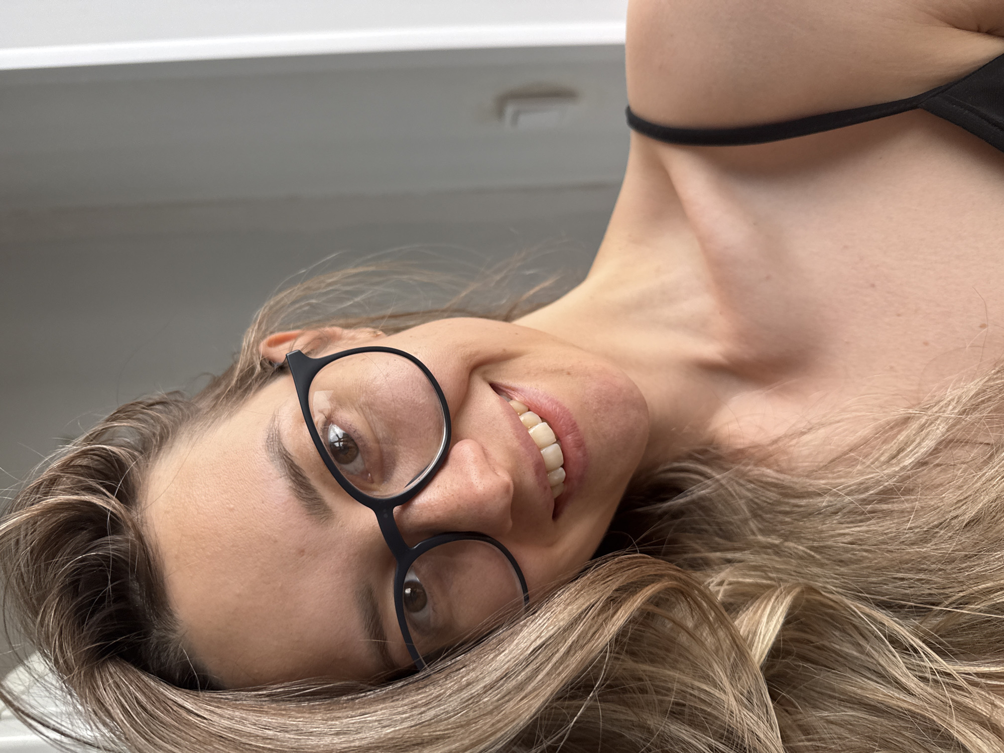 The image displays a woman from a close-up perspective, with the photograph rotated roughly 90 degrees counter-clockwise. She is smiling broadly and looking directly at the viewer. Her head is positioned on the left side of the frame, with her body extending to the right. She has light brown to blonde, wavy hair that partially covers her shoulders and chest.
She is wearing black-framed, round glasses, and her brown eyes are clearly visible behind the lenses. Her smile is natural and wide, showing her upper teeth. A black strap, likely from a top or swimsuit, is visible across her upper chest and shoulder area. The background is a plain, light grey surface, which could be an interior wall or ceiling, suggesting an indoor setting. The lighting is soft and even. No text is visible within the image. Her expression conveys a sense of happiness and engagement. While the image itself does not offer specific geographical indicators, it is contextualized as being from Chaikovsky, Russia.