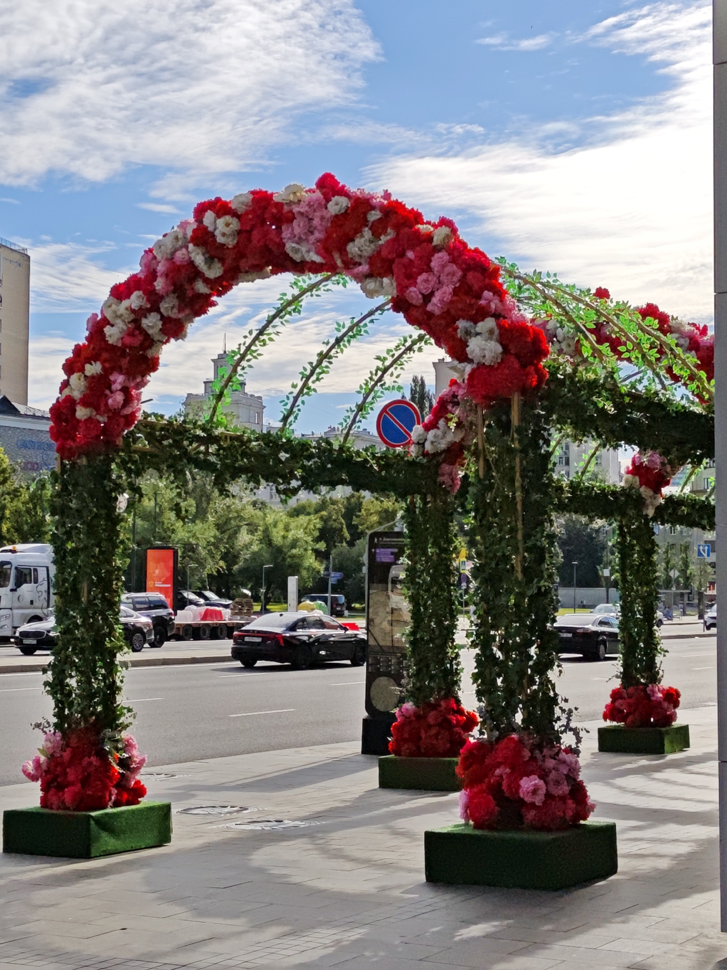 This image depicts an outdoor decorative archway adorned with abundant red and pink artificial flowers and green ivy. The archway stands on a paved pedestrian walkway, suggesting a public space, likely a street or plaza in Moscow, Russia. Beyond the arch, a road with several cars is visible, including a black sedan in the foreground. A white truck and other vehicles are further down the road, indicating moderate traffic. In the background, buildings and trees line the street, under a bright blue sky with scattered white clouds, suggesting a pleasant, possibly sunny day.
The archway is a prominent feature, suggesting an event or celebration is taking place or has recently occurred. Its elaborate floral decoration points towards a festive or welcoming atmosphere. The presence of cars and buildings provides context for an urban environment. There is a circular blue and red sign on a pole, possibly a traffic sign, visible behind the arch. Several green rectangular bases, topped with similar floral arrangements, are placed along the walkway in front of the arch. The image is captured from a slightly low angle, emphasizing the grandeur of the archway against the sky. No people are clearly visible in the frame.