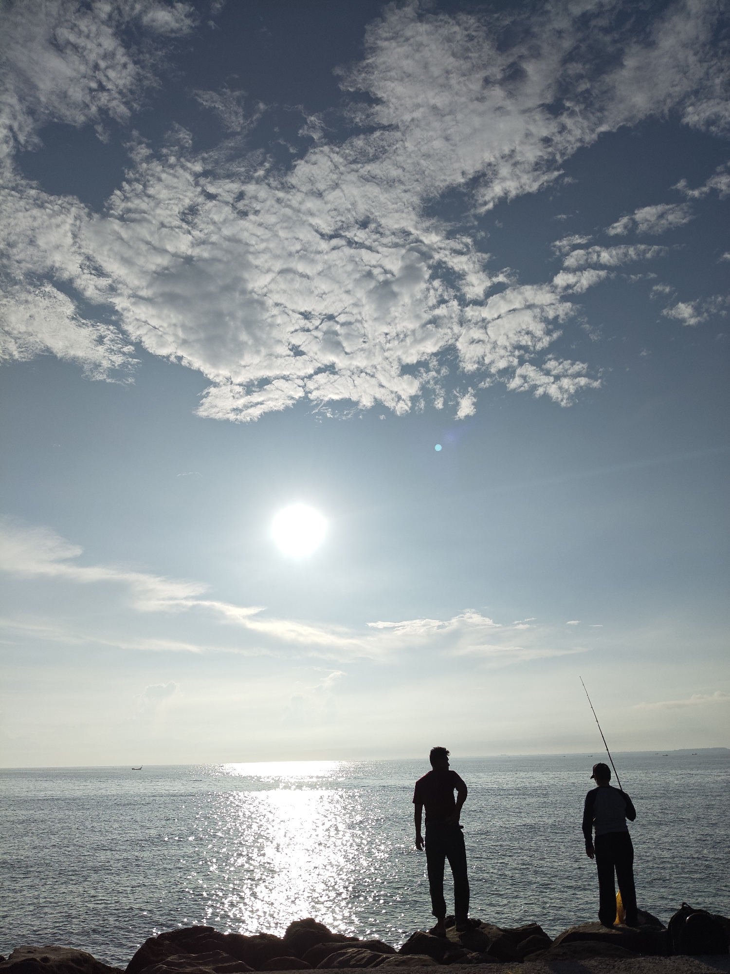 The image depicts a coastal scene in Lhokseumawe, Indonesia. Two silhouetted figures stand on a rocky shore, likely fishing. One holds a fishing rod. The sea stretches out to the horizon, shimmering with reflected sunlight. In the distance, a small boat is visible. The sky is partly cloudy, with the sun positioned centrally, creating a strong glare. The environment is bright and sunny.