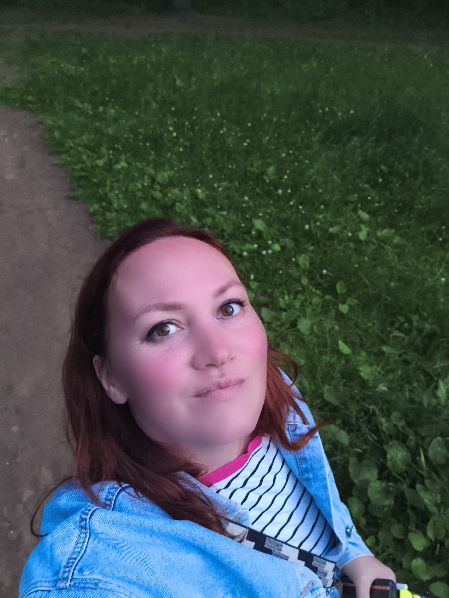 A woman with long, auburn hair is captured in a close-up, upward-angled selfie outdoors. She is wearing a bright blue denim jacket over a striped t-shirt with black and white horizontal lines and a pink neckline. Her left shoulder and part of her arm are visible. Her face is slightly turned, with her gaze directed upwards and to the left, and a gentle smile playing on her lips. Her cheeks have a noticeable pink blush.
The background consists of a grassy area with scattered white flowers, suggesting a natural setting like a park or meadow. To the left of the frame, a dirt path curves away, bordered by the grass. The lighting indicates it is likely late afternoon or early evening, with soft, diffused light. The overall mood is relaxed and pleasant. There is no visible text in the image. The location context is Chaikovsky, Russia.
