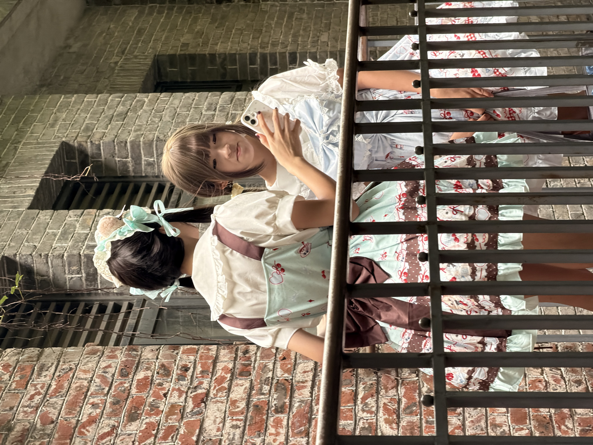 The image shows two young women dressed in Lolita-style dresses. One woman is in the foreground, with her back to the camera, wearing a light-colored dress with a mint green apron featuring a heart and bear pattern. She has dark hair with a light blue bow and what appears to be a cat-ear headband. The other woman is in the background, facing the camera, wearing a white blouse with ruffled sleeves and a light blue dress with a white lace trim. She is holding a smartphone in her right hand and looking at it. Both women are standing on a metal walkway, possibly a balcony or bridge, with horizontal bars in the foreground and background. The setting appears to be outdoors with brick walls visible behind the metal walkway. The lighting suggests it might be daytime, possibly late afternoon or early evening, given the somewhat diffused light. The overall mood appears casual and relaxed, with the woman in front taking a selfie or video. There is no visible text in the image.