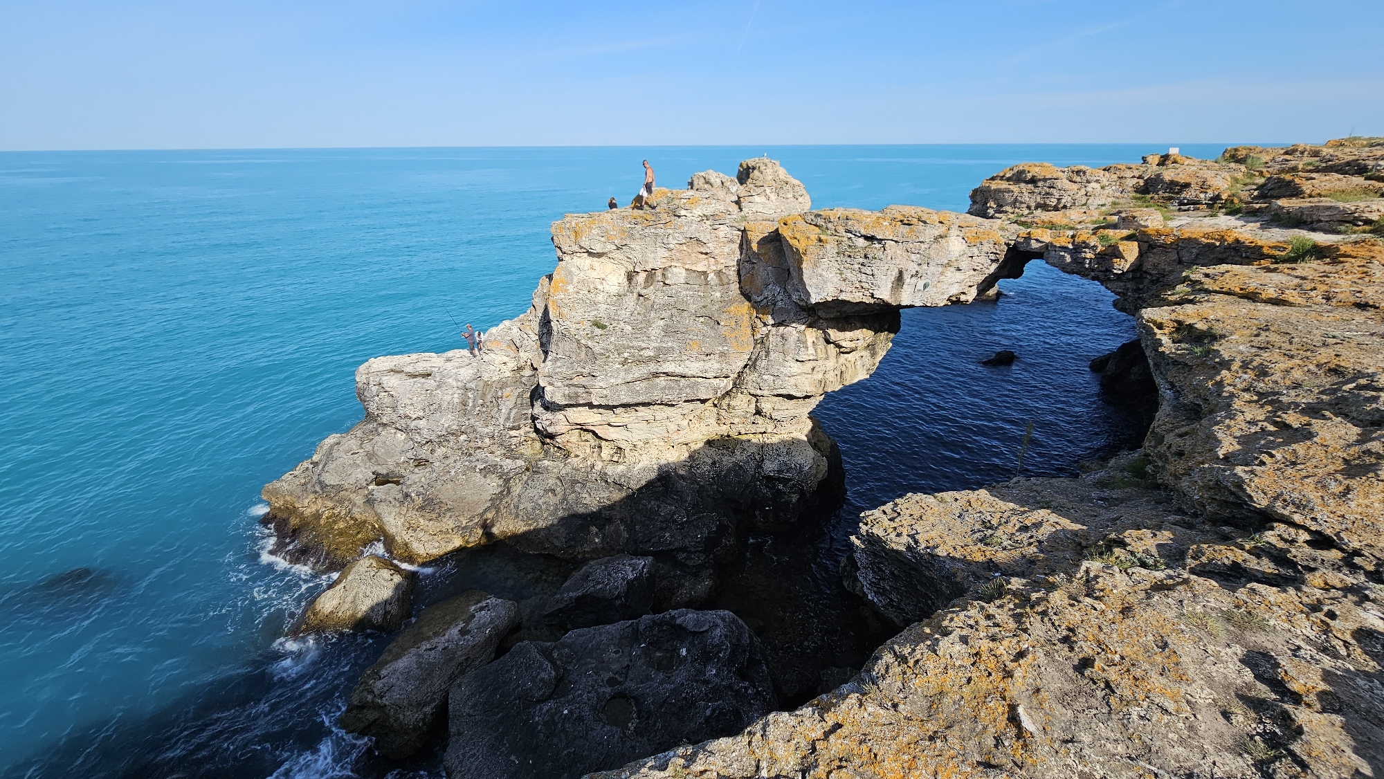 The image shows a natural rock arch formation on the coast of Unknown, Unknown. The arch spans a body of water, connecting a large rock outcropping on the left with a higher, rocky cliff on the right. The water is a vibrant turquoise color. There are three people visible on the rock formation: two on the left-hand side of the arch, one possibly fishing, and another person standing on top of the arch. The sky is clear and blue. The rock formations are layered and show signs of weathering and orange lichen growth. The angle of the shot is from above, looking down on the arch and the water.