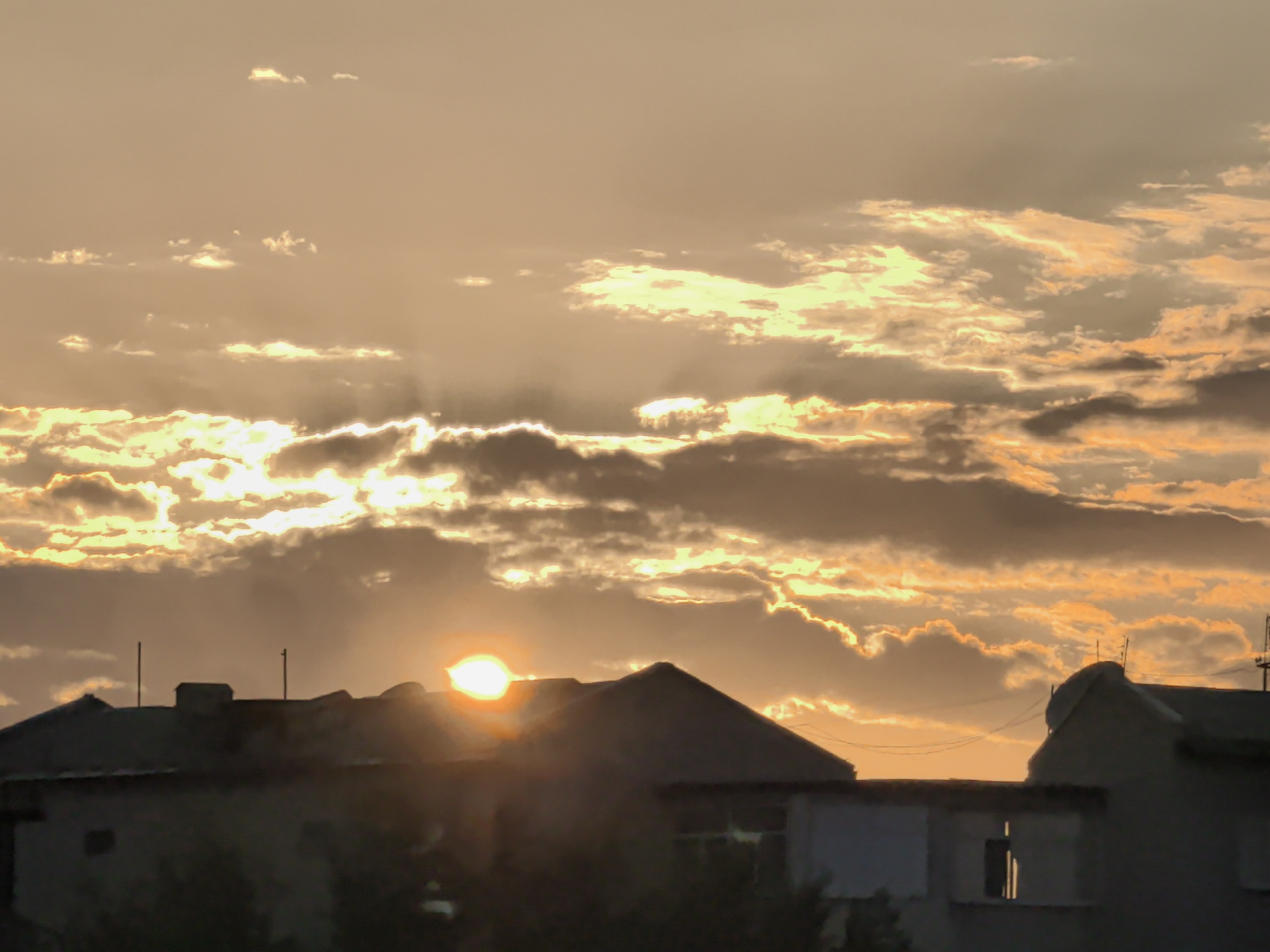 The image captures a sunset over a residential area, likely in Taldykorgan, Kazakhstan. Silhouetted buildings, appearing to be houses with pitched roofs, dominate the foreground. Their dark forms contrast with the vibrant sky. Two thin, vertical poles, possibly antennae or support structures, are visible on the rooftops.
The sky is filled with dramatic clouds, illuminated by the setting sun. Rays of light break through the clouds, creating a golden and orange glow. The sun itself is a bright, orb-like shape, partially obscured by a cloud on the right side of the frame, casting a warm light. The overall impression is one of a serene and picturesque sunset. The time of day is clearly evening, as indicated by the sunset. The weather appears to be clear enough for a visible sunset, though clouds are present. There is no visible text in the image.