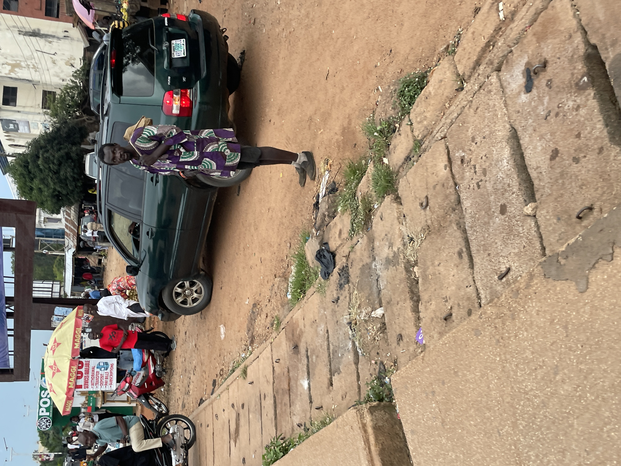 The image depicts a street scene in Jos, Nigeria. In the foreground, a man stands on a dirt road next to a dark green SUV. He is wearing a purple and white patterned traditional garment and dark shorts, with a bag slung over his shoulder. The SUV has a Nigerian license plate that reads "KRD 589CO". Behind the man and the SUV, several other people are visible, as well as a red motorcycle and an umbrella stall with "MAGGI" branding. The environment appears somewhat dusty, with scattered debris on the ground. The buildings in the background suggest an urban setting.