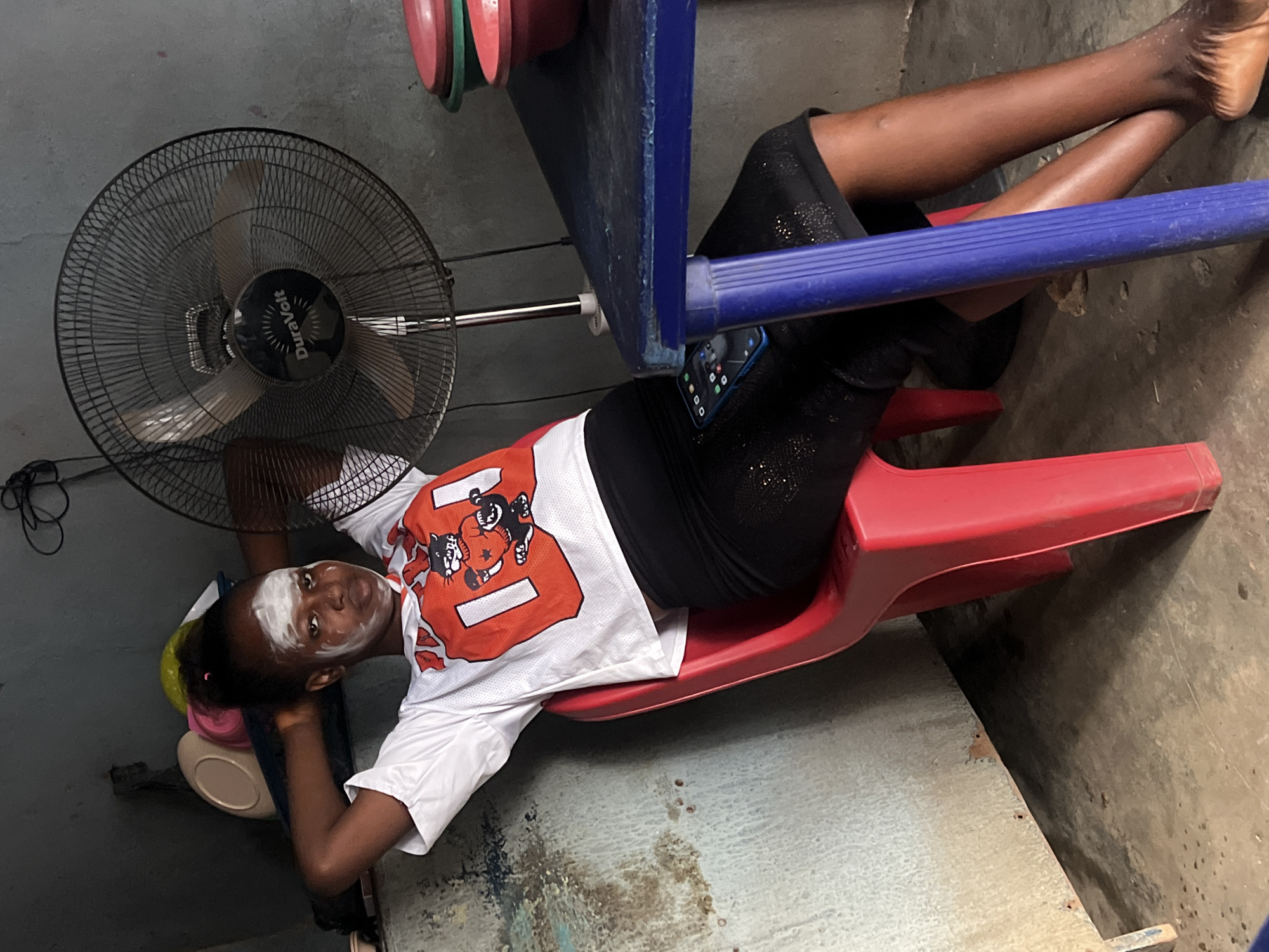 In Nkpor, Nigeria, a young woman with white cream on her face sits on a red plastic chair. She wears a white t-shirt with an orange graphic of a cartoon cat and a black skirt. Her hands are clasped behind her head. A black oscillating fan with the brand "DuraVolt" is visible behind her. In front of her, to the right, is a blue table with several colorful bowls on it, and a cell phone is placed on her lap. The environment appears to be indoors, with a concrete floor and a textured wall.