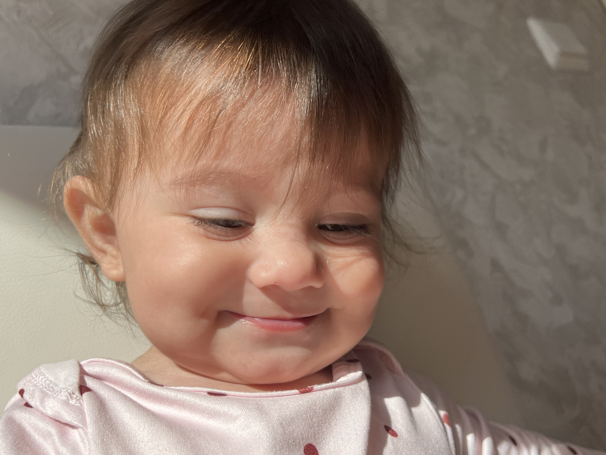 A close-up shot captures a baby, likely between 6 and 18 months old, with fair skin and brown hair, smiling slightly while looking downward. The baby is wearing a pink garment with dark red polka dots. Sunlight casts shadows across the baby's face and upper body. The background appears to be a plain, neutral-colored wall. There is no evidence suggesting the location is Elena, Bulgaria.