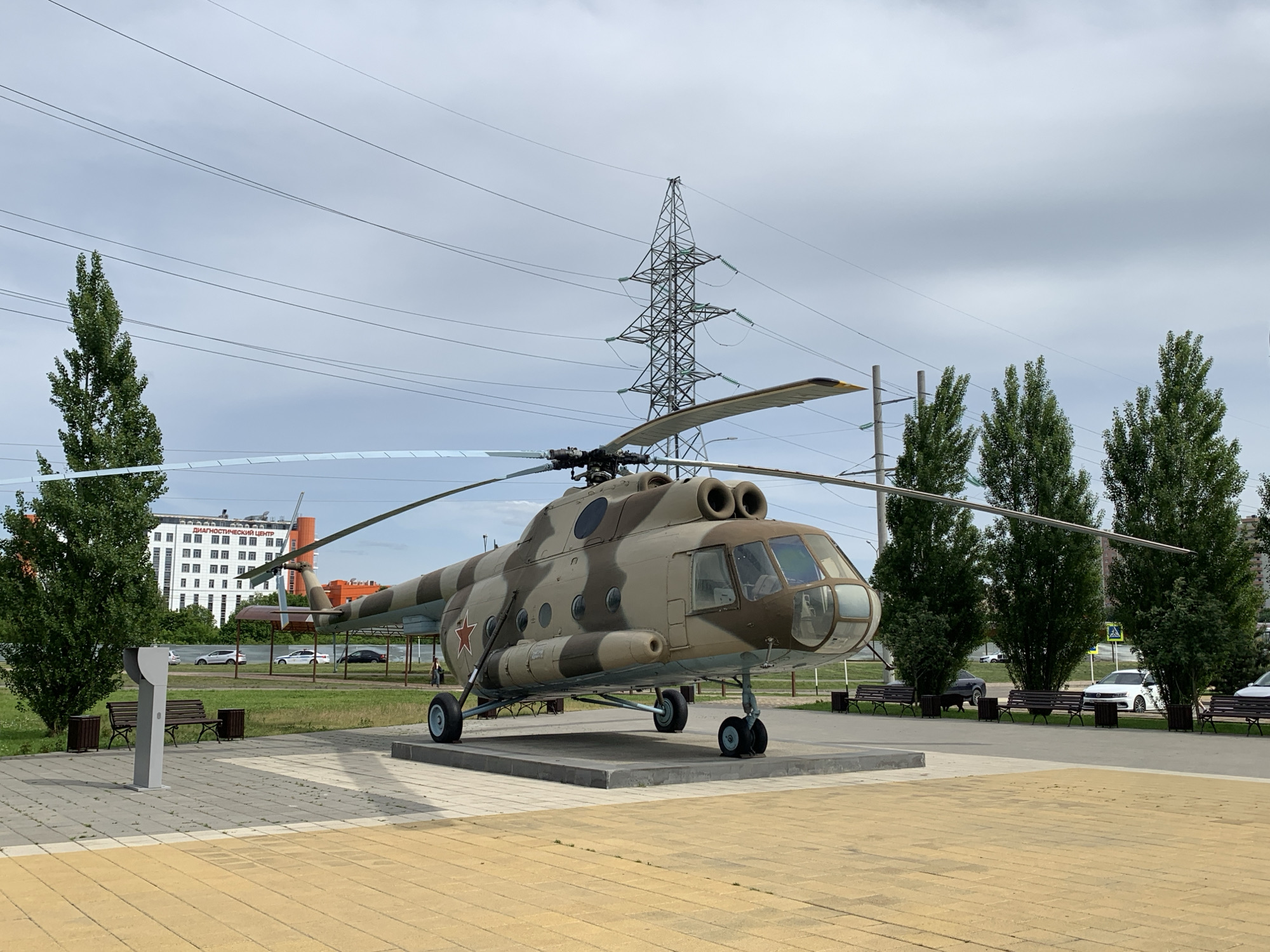 An outdoor, wide-angle, eye-level, daytime shot depicts a military helicopter on display, positioned on a concrete platform. The helicopter, camouflaged in brown and tan, has its rotor blades extended. Behind the helicopter, a tall, lattice-style electrical tower with multiple wires stretching across the sky is visible, along with several tall, green trees. In the background, a white building with many windows, identified by signage as a "DIAGNOSTIC CENTER," stands prominently. The foreground features a yellow, brick-paved area and a gray walkway with benches and trash receptacles. Several cars are parked along a road bordering the display area, and a few people are present in the scene. The sky is overcast with clouds.