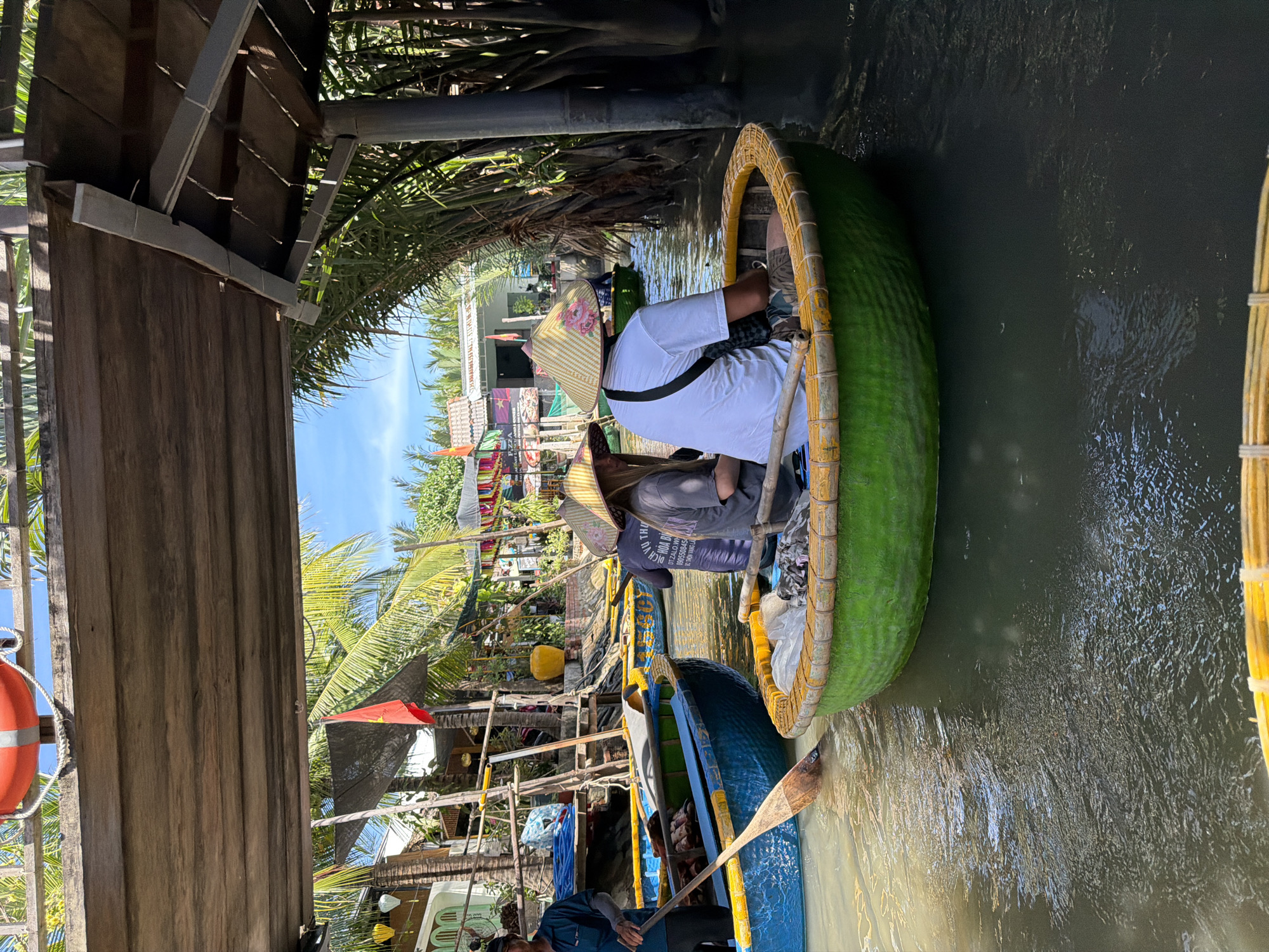 In a canal in Da Nang, Vietnam, several circular basket boats are navigating under a wooden pier. The scene is illuminated by daylight, with a clear blue sky visible. In the foreground, a green basket boat contains two individuals wearing conical hats, one of whom is rowing. Another blue basket boat is to the left, with a person in a dark shirt and hat on the left side, holding a wooden oar in the water. In the background, more basket boats are visible, as well as buildings with palm trees and a Vietnamese flag. The water in the canal appears murky.