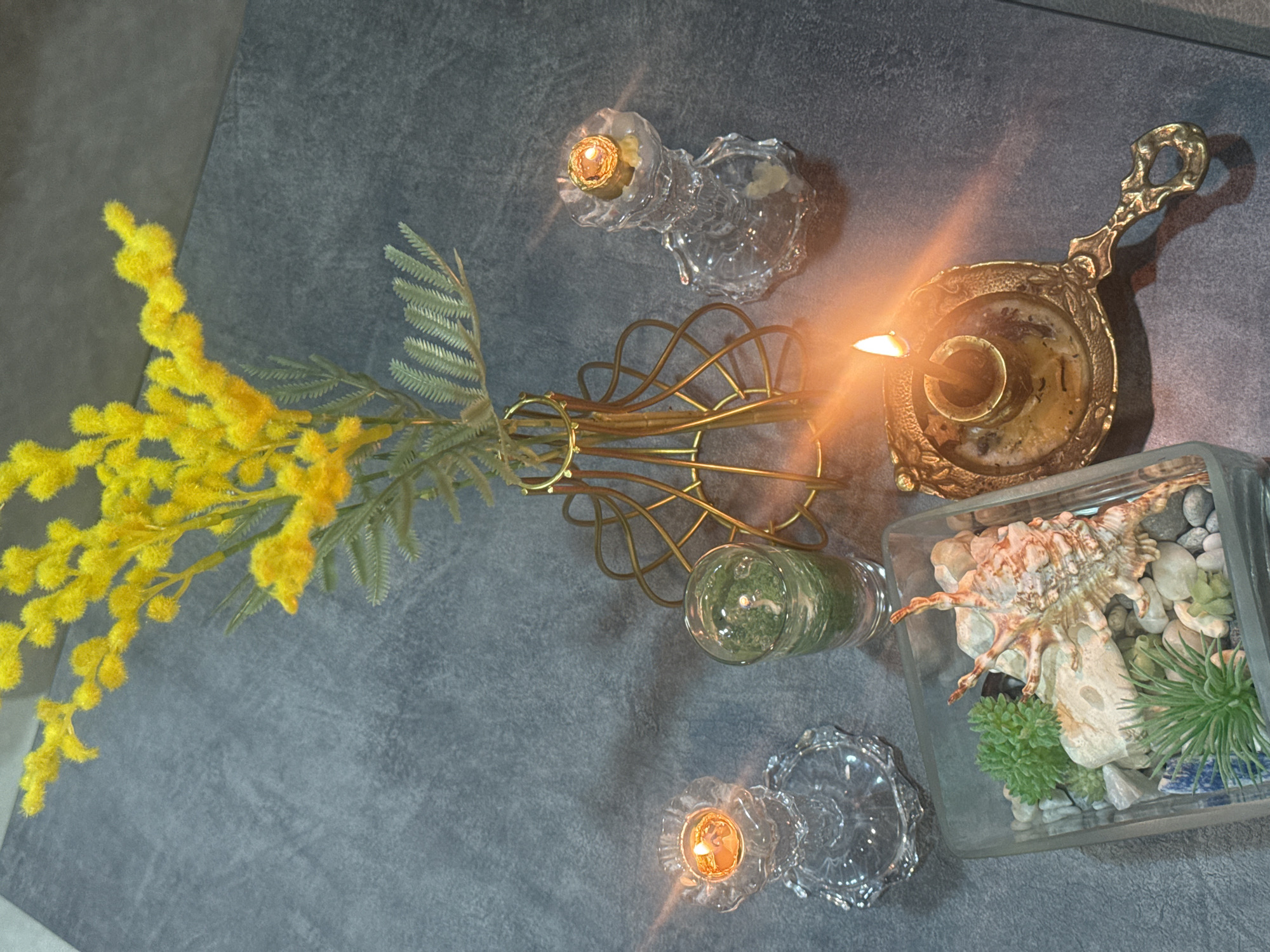 A top-down view shows a still life arrangement on a gray surface. A vibrant yellow Mimosa branch with feathery green foliage is positioned to the left, emerging from a gold-colored wire vase. Two small, clear glass candle holders with lit candles are placed on either side of the arrangement. A vintage gold candle holder with a lit candle sits near a square glass container filled with white stones, faux green plants, and a large seashell.
