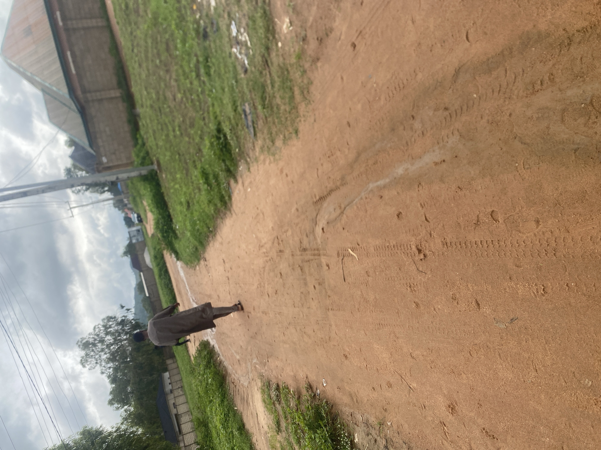 A lone individual walks away from the camera on a dirt road in Howayi, Nigeria. The person is wearing a long, loose-fitting, dark grey garment and sandals. The road is reddish-brown and appears to be a well-traveled path, with visible tire tracks and footprints. To the left of the road, there is lush green grass and some vegetation, with fences and buildings visible in the background. To the right, the grass is also green, with some scattered debris and more buildings, including structures with distinct angled roofs. The sky is overcast with heavy clouds, suggesting a cloudy or potentially rainy day. Power lines are visible stretching across the upper portion of the frame. The overall scene depicts a rural or semi-urban setting within Howayi.