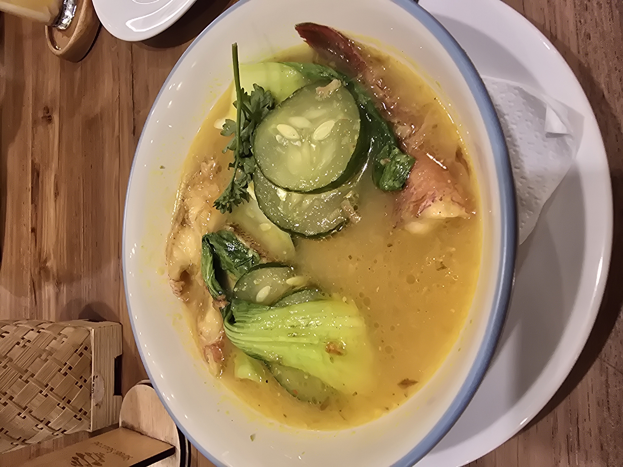 A high-angle shot shows a bowl of soup, seemingly on a wooden table, possibly in a restaurant setting in Nusa Penida, Indonesia. The soup, with a yellow broth, contains visible ingredients such as sliced cucumbers, green vegetables that might be bok choy, and shrimp. The bowl is white with a blue rim and sits on a white plate with a folded white napkin. Adjacent to the bowl is a woven box with a wooden stand. Other objects, including a drink, a small white plate, and the edge of the table, are also visible. The setting suggests an informal meal in a casual dining atmosphere.