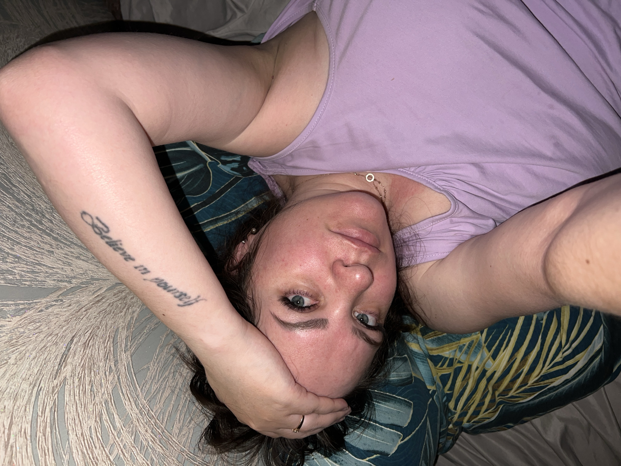 A woman lies on her back on a bed. She wears a light purple tank top and a gold necklace. Her right arm is raised, with the hand touching her forehead. A tattoo that reads "Believe in yourself" is visible on her right forearm. The bed has a gray textured sheet and a pillow with a blue and gold tropical leaf pattern. The location is unknown and there is no indication of it being Noviy, Russia.
