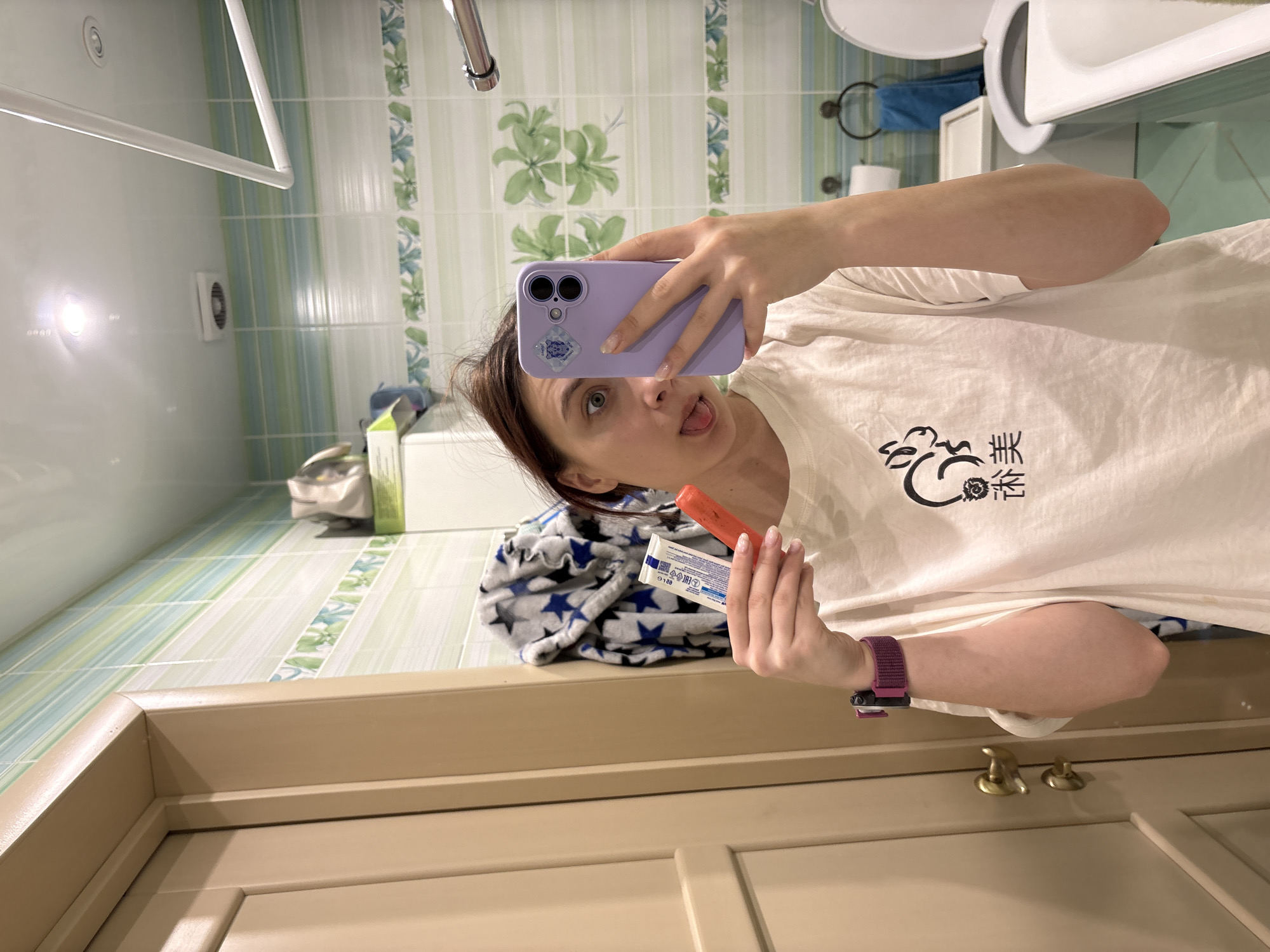 A person is taking a selfie in a bathroom with a purple iPhone. The person is holding a lipstick and its packaging in their right hand, and their tongue is slightly sticking out. They are wearing a white t-shirt with a black graphic.
The bathroom has light green and white striped tiled walls with a floral border. A toilet is visible in the background. The floor is tiled in a similar style. There is a door in the foreground.
