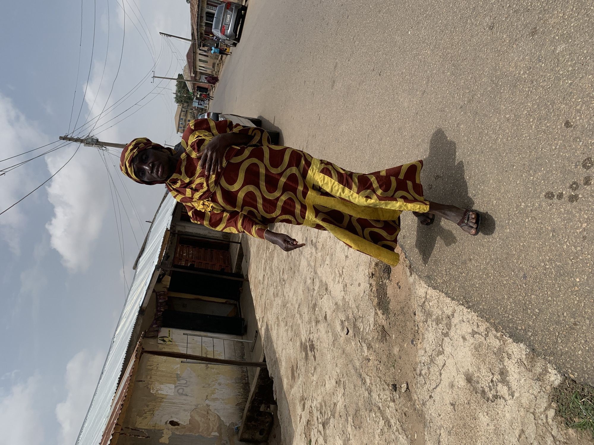 A woman stands in the street in a town outside of Jos, Nigeria. The woman is wearing a long, patterned dress of red and yellow, and a matching head covering. She has on sandals. The road is grey asphalt and the sky is cloudy. A building, possibly a shop, is on the left. A car is parked further up the road and there are people in the background. There are telephone poles with wires running overhead.