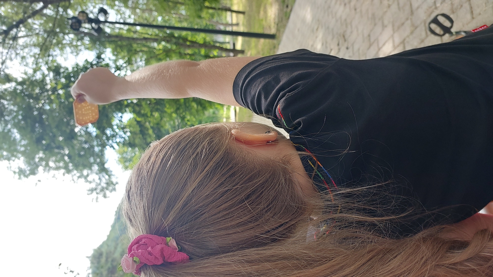 A young girl, seen from behind, stands outdoors in Lermontov, Russia, holding a cracker with small holes in it aloft. She has long, blonde hair pulled back with a pink scrunchie and wears a black t-shirt with colorful embroidery. The background features green trees, a paved walkway, and a bright sky. A bicycle pedal is visible in the lower right corner.