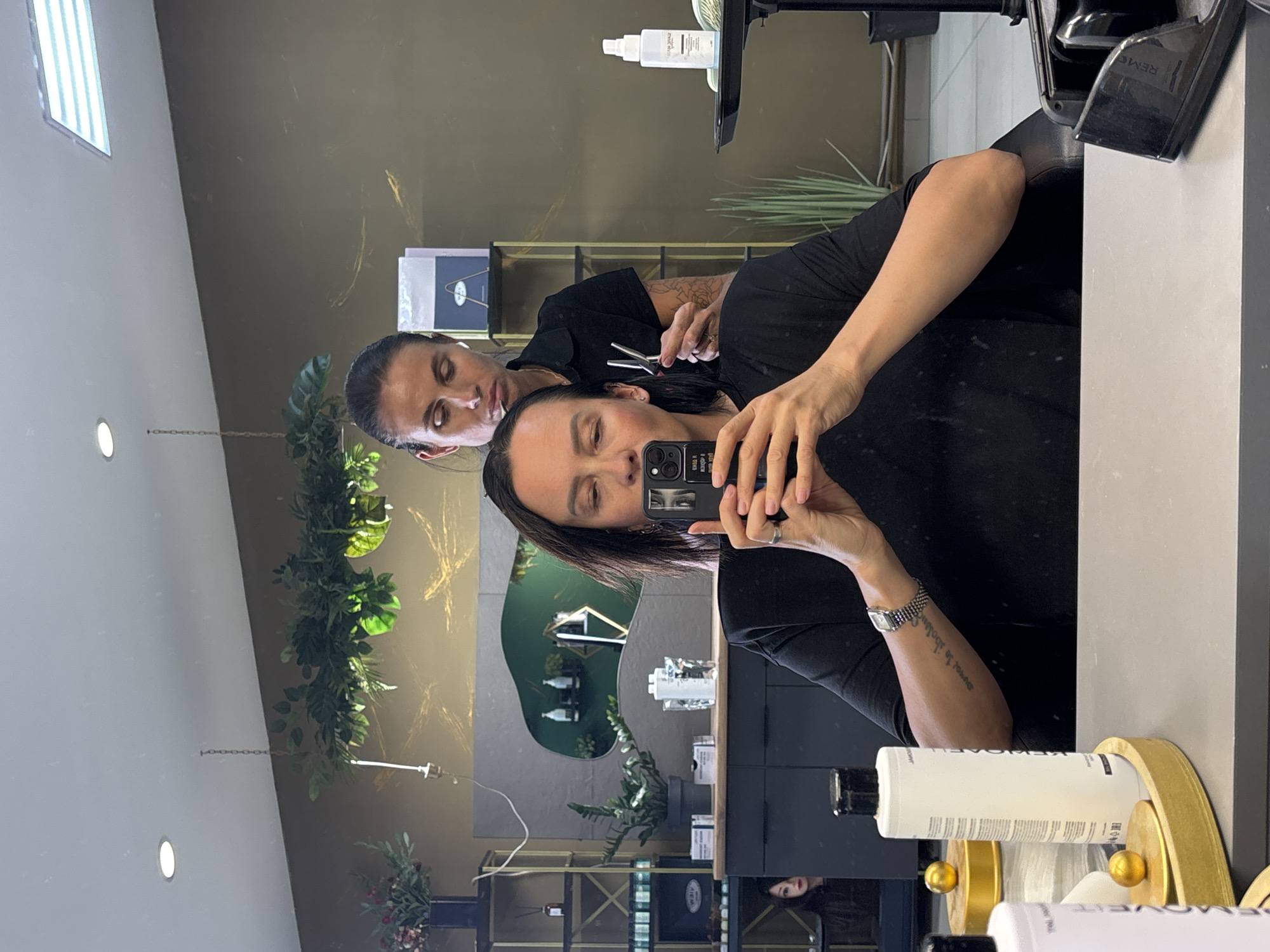 A mirrored perspective of two individuals inside what seems to be a hair salon. One person, holding a black smartphone, appears to be taking a selfie with their reflection. The other person, positioned behind and to the left, is holding a pair of scissors, seemingly in the act of cutting or styling hair. The setting includes salon equipment and products, decorative plants, and shelving with various items. There's a neutral-toned wall visible with gold accents. The exact location of the image is not explicit in the image, but as you specified Chaikovsky, Russia, it would be a hair salon in that city.