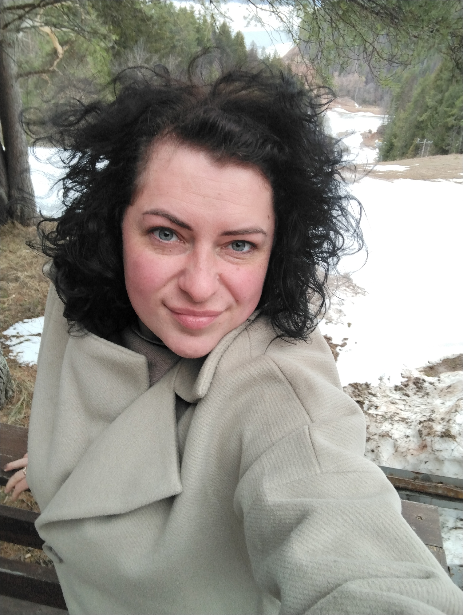 A woman is pictured outdoors, possibly in Galevo, Russia. She has dark, curly hair and blue eyes. She is wearing a light-colored coat. The background shows a snowy landscape with trees. She is sitting on a wooden bench.