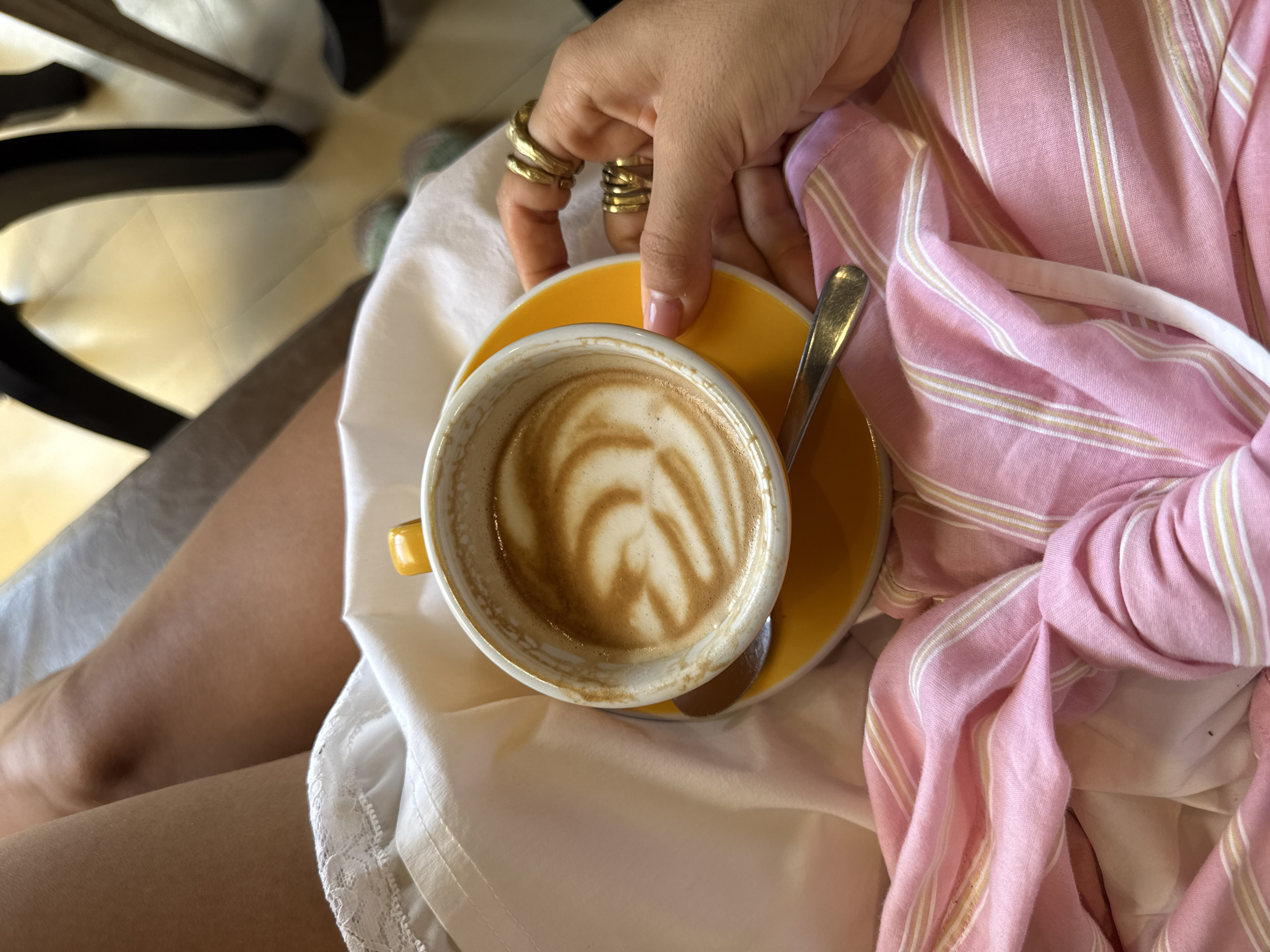 A high-angle shot captures a woman's hands and legs holding a yellow cup and saucer of coffee. The coffee has a latte art pattern. The woman wears several rings on her fingers. She's wearing a white dress with lace detailing and a pink and white striped shirt. The scene takes place indoors, the leg of a black chair visible in the top left. The location appears to be in North Kuta, Indonesia.