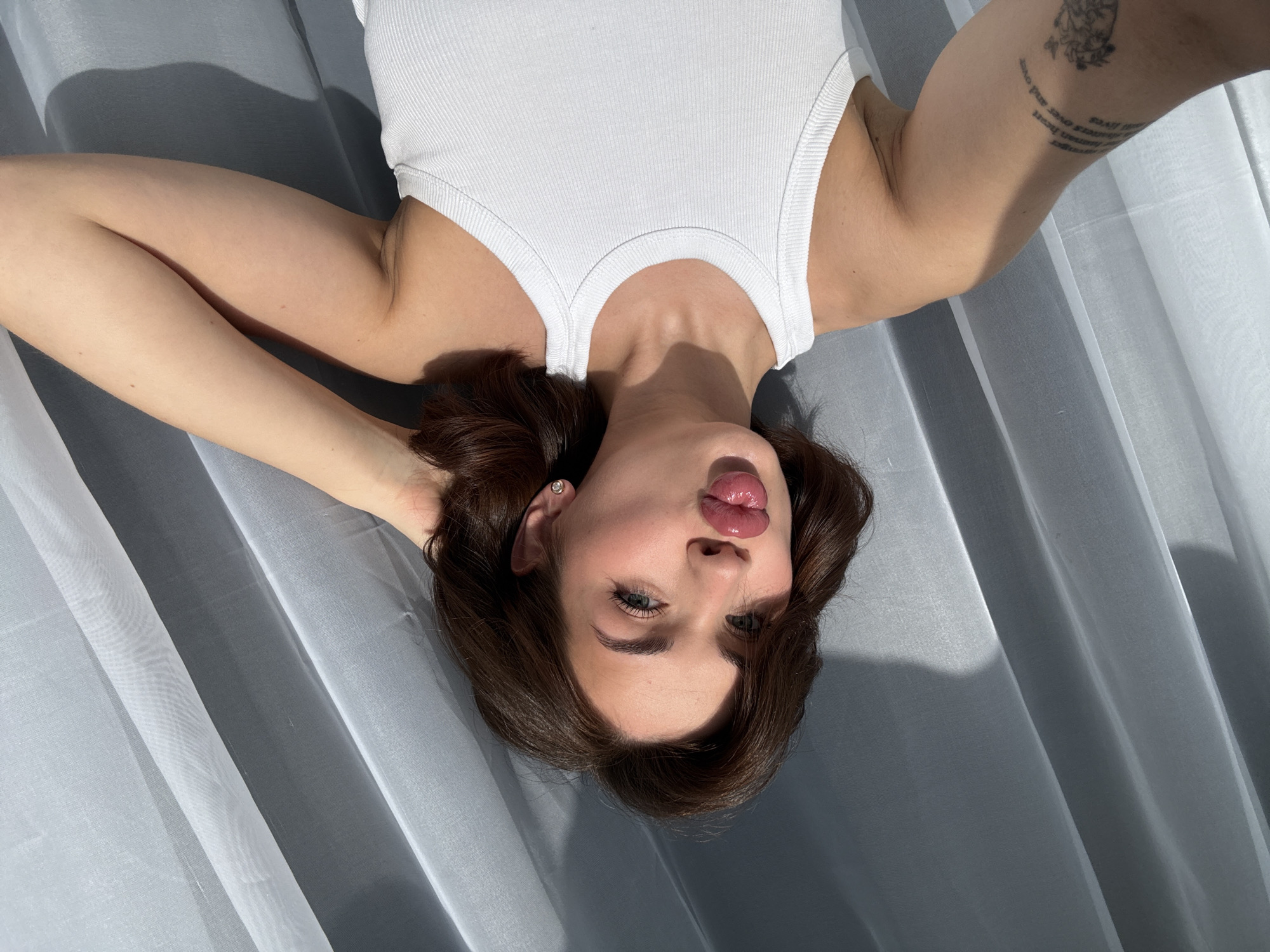 A person with light skin, dark brown hair, and light eyes is lying on a light gray fabric surface, possibly a curtain or draped sheet. The person is wearing a white tank top. The person is facing the camera with their head tilted upside down, pursing their lips slightly. Their arms are raised above their head, with a tattoo visible on their right forearm. The lighting is bright and diffused, creating shadows on the fabric. The background is plain and out of focus, consisting of the same light gray fabric. There is no indication that this photo was taken in Sarapul, Russia.