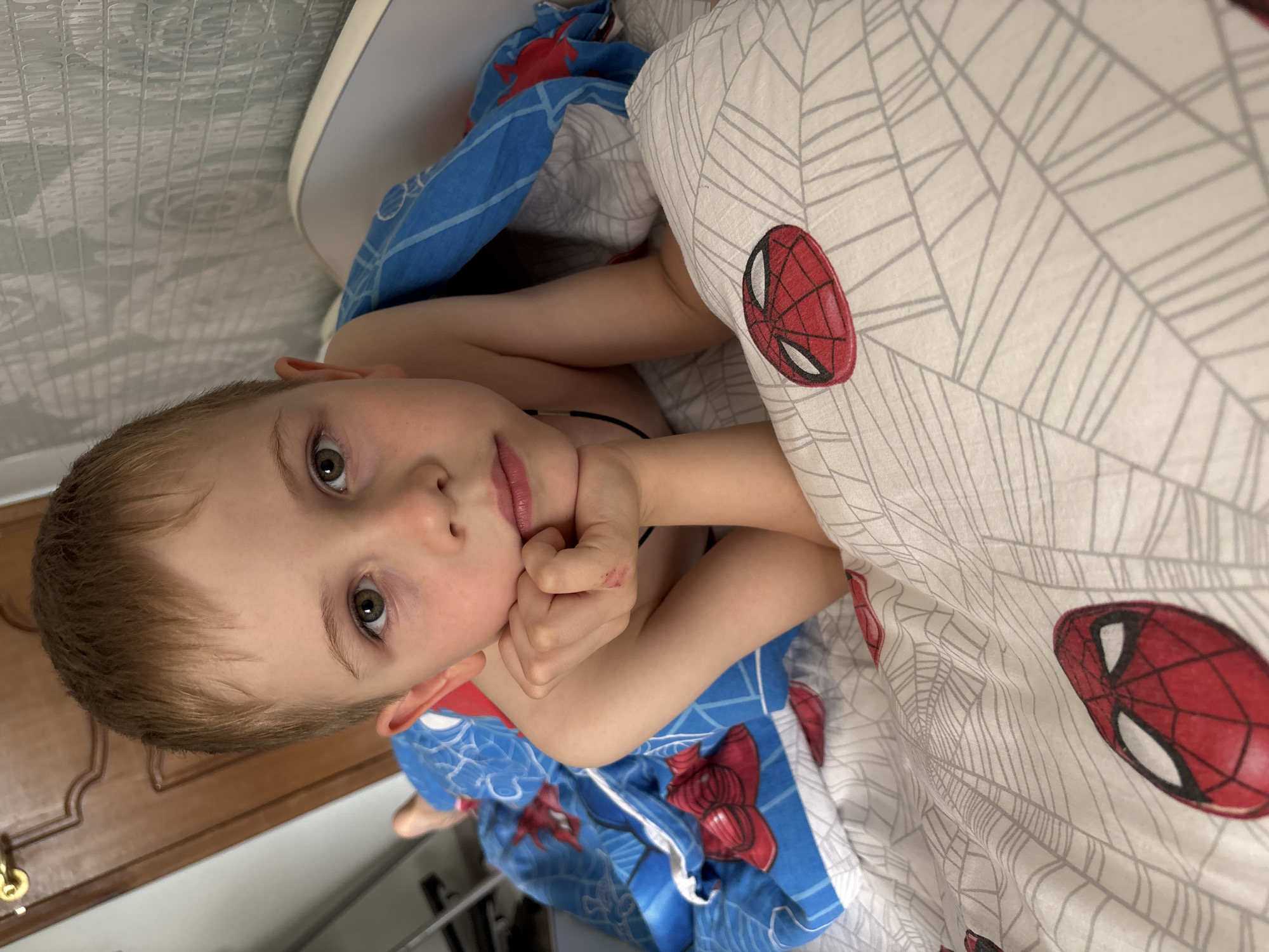 A young boy with light brown hair and green eyes is lying in bed, looking up at the camera. He is partially covered by a blue blanket and a white duvet with a Spider-Man theme. The boy's right hand is near his face, and he appears to be resting. The lighting suggests it could be daytime. The setting appears to be a bedroom, with a wooden wardrobe visible in the background. The duvet has a repeating pattern of Spider-Man masks and spider webs. The boy's expression is calm and perhaps a little sleepy.