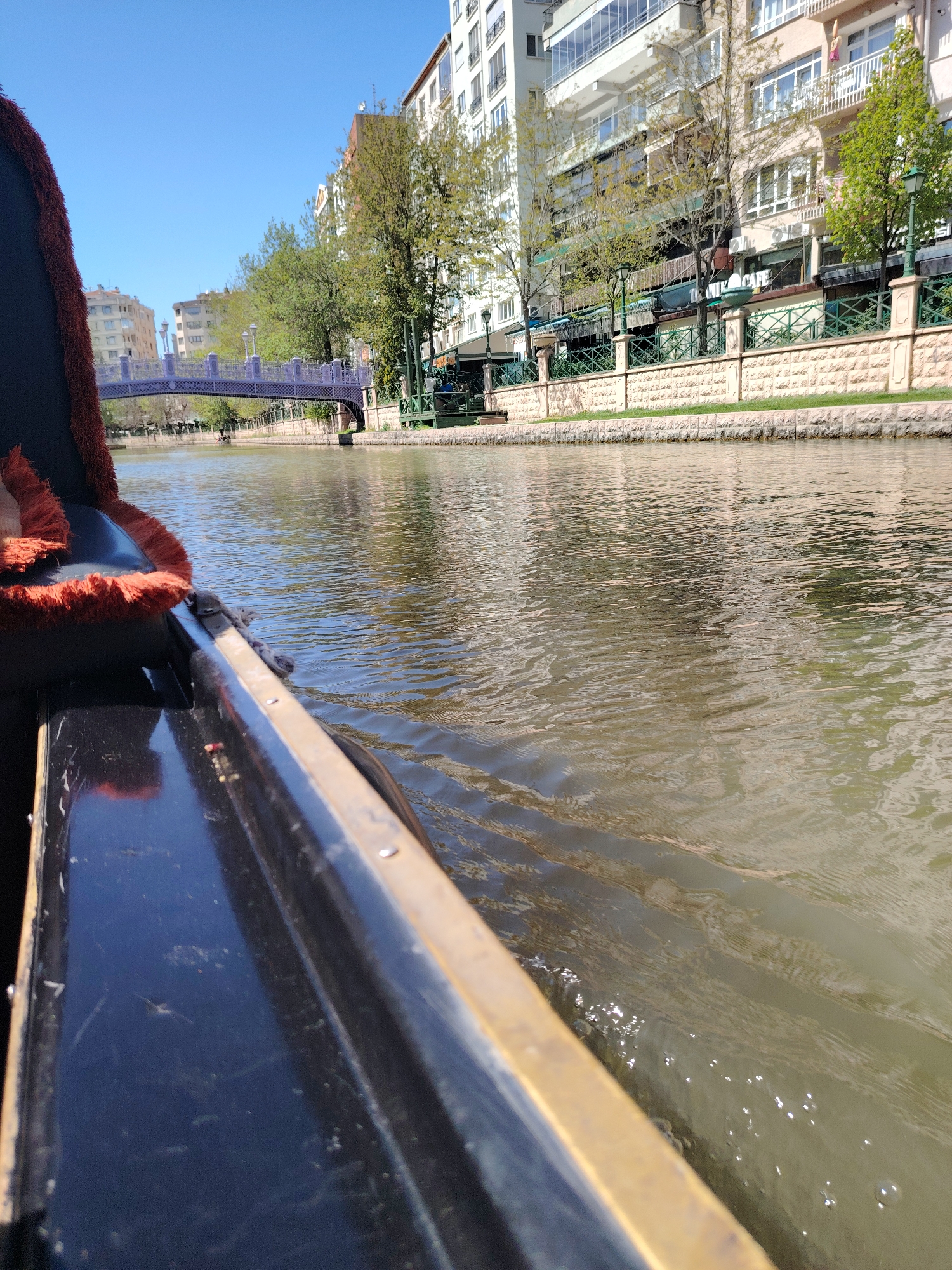 The image provides a first-person perspective from a boat, likely a gondola, cruising down a river or canal in Eskisehir, Turkey, on a bright, sunny day. The left side of the frame shows the dark, possibly black, hull of the boat with a prominent golden-colored rim. A reddish-orange, textured cushion, part of the boat's seating, is visible on the far left.
The water, occupying a significant portion of the image, displays ripples and reflections of the clear blue sky and surrounding buildings, indicating the boat is in motion. The right bank of the waterway features a promenade lined with multi-story buildings, characterized by balconies and numerous windows. Interspersed among the buildings are mature green trees. A decorative stone wall with green railings and pillars runs along the bank. Visible on one of the buildings is a sign that partially reads "TITANIK CAFE."
In the mid-ground, a light purple or blue arch bridge spans the river, connecting the two sides. Beyond the bridge, more urban structures and foliage are visible. The sky is clear and blue, suggesting pleasant weather conditions. The overall scene depicts a serene and leisurely boat ride through an urban landscape.