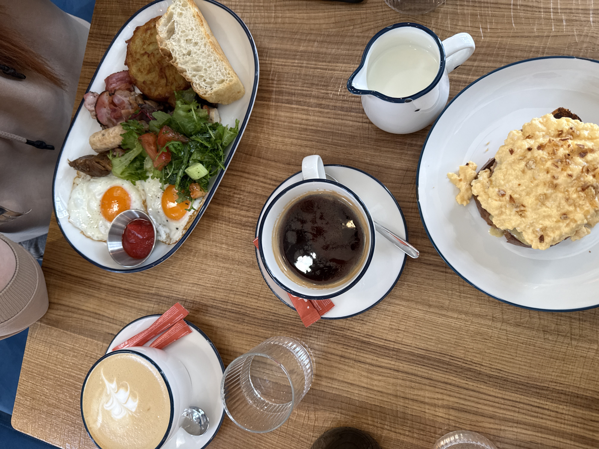 The image depicts a top-down view of a wooden table set with a breakfast meal, likely taken in Krasnoyarsk, Russia, based on the style of food and drink.
On the left, a white plate with a blue rim contains a full English breakfast: two fried eggs with runny yolks, a small metal container of ketchup, several pieces of sausage and bacon, a fried potato cake, a slice of bread, and a mixed green salad with tomato and cucumber. Next to this plate, a fair-skinned person's upper body, including their shoulder and a beige garment, is partially visible.
To the right, a white pitcher with a blue rim is filled with milk or cream. Adjacent to this is a white saucer with a blue rim holding a white coffee cup, also with a blue rim, filled with dark coffee. A metal spoon rests on the saucer. Two red sugar packets sit beside the cup.
In the lower left corner, a white coffee cup with a blue rim on a matching saucer contains a latte with latte art. A small metal spoon lies beside the cup. Next to this is a clear ribbed glass, likely empty.
Further to the right, a white plate with a blue rim holds a serving of scrambled eggs on toast. The wooden table has a visible grain pattern. Two unidentified objects are at the bottom right.