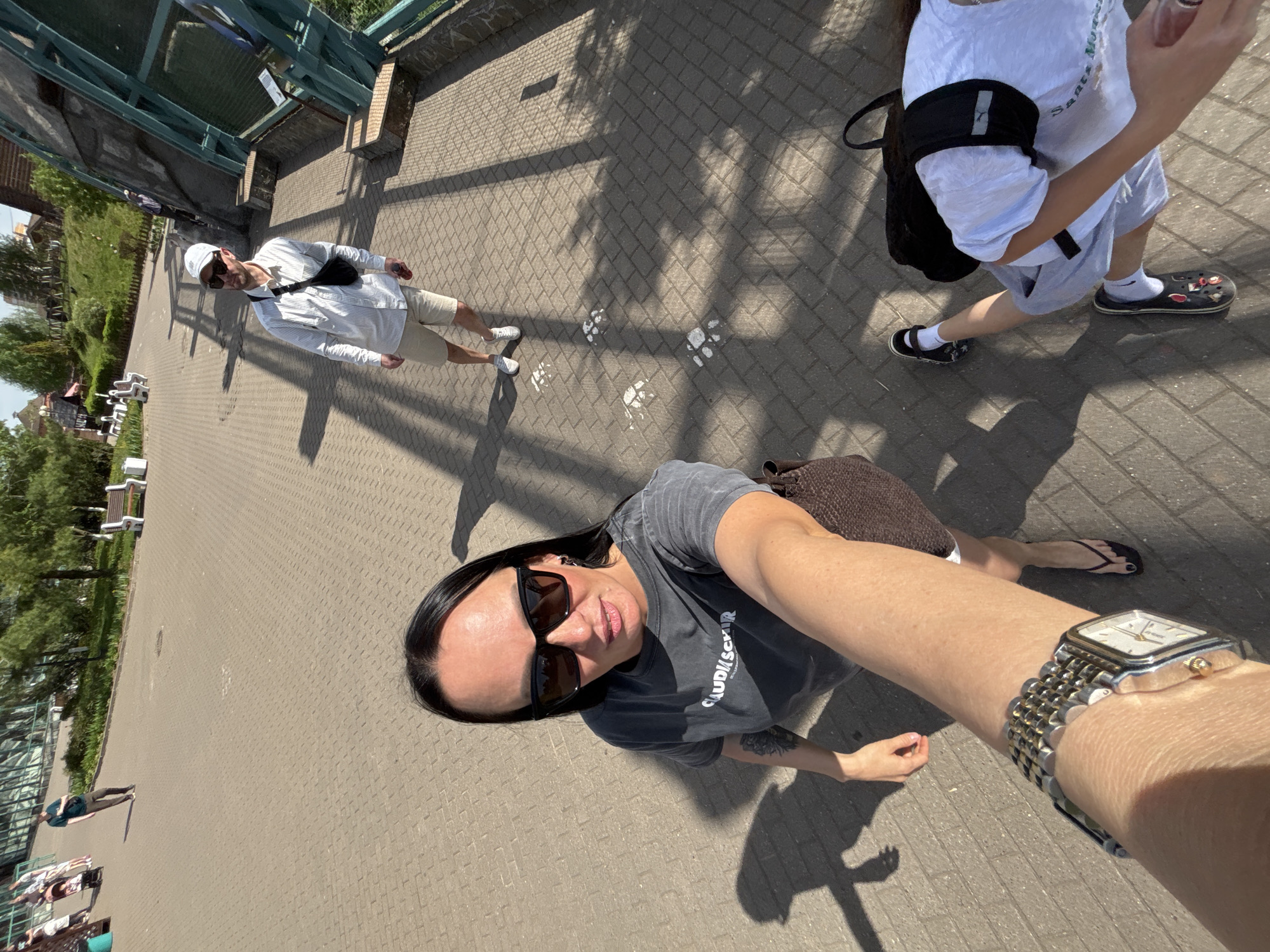 The image, taken from an elevated perspective, features a woman holding the camera out to capture a selfie. She is wearing sunglasses, a grey t-shirt, and a watch on her left wrist. Two other individuals are visible: an adult male in a white shirt and light-colored shorts and a child in a white shirt and light-colored shorts. The setting appears to be an outdoor pedestrian area with a paved surface, possibly a park or public space. White animal footprints are visible on the pavement. Greenery, including trees and landscaping, can be seen in the background, as well as benches, possibly in a seating area. Some sort of glass or metal structure is to the left of the image. Based on the prompt, the location is in Izhevsk, Russia.