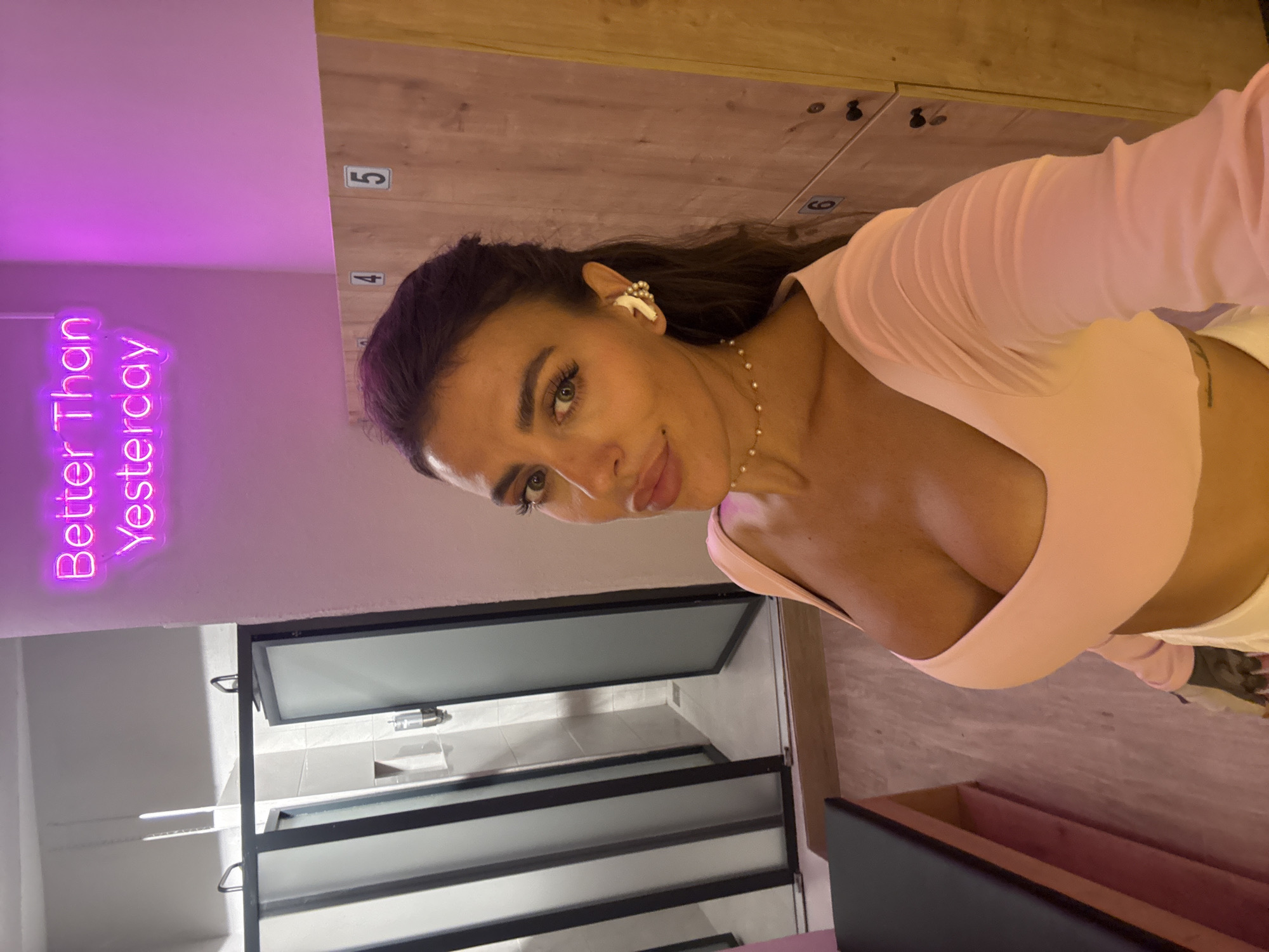 A woman poses for a selfie in a locker room. She is wearing a light pink top and white pants. Her hair is dark and pulled back, and she has on white earbuds. A pearl necklace is visible around her neck. In the background, wooden lockers are stacked, with numbers 4, 5, and 6 visible on them. To the left of the woman, a neon sign reads "Better Than Yesterday" in bright pink. The scene appears to be indoors, possibly a gym or spa, with a grey tiled floor and frosted glass doors visible. The lighting is dim with pink and purple tones from the neon sign. The woman's expression is a slight smile. There are no clear indications of the time of day or weather, but the indoor setting suggests it's not dependent on external conditions. The location cue of Fethiye, Turkey, provides a geographical context.