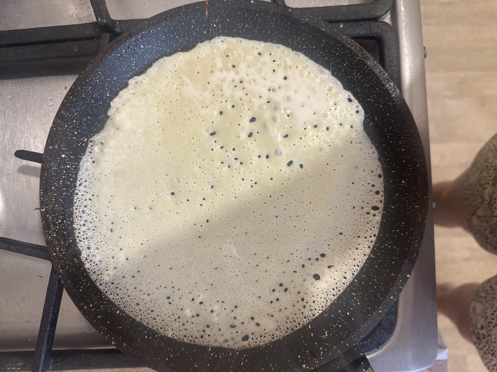 A pancake is being cooked in a frying pan on a stovetop. The pancake is light in color and covered in small bubbles and darker spots. The frying pan is black with a speckled pattern and is resting on a black stovetop burner. A person's leg and bare foot are partially visible on the right side of the image. There is no information available that indicates this photo was taken in Votkinsk, Russia.